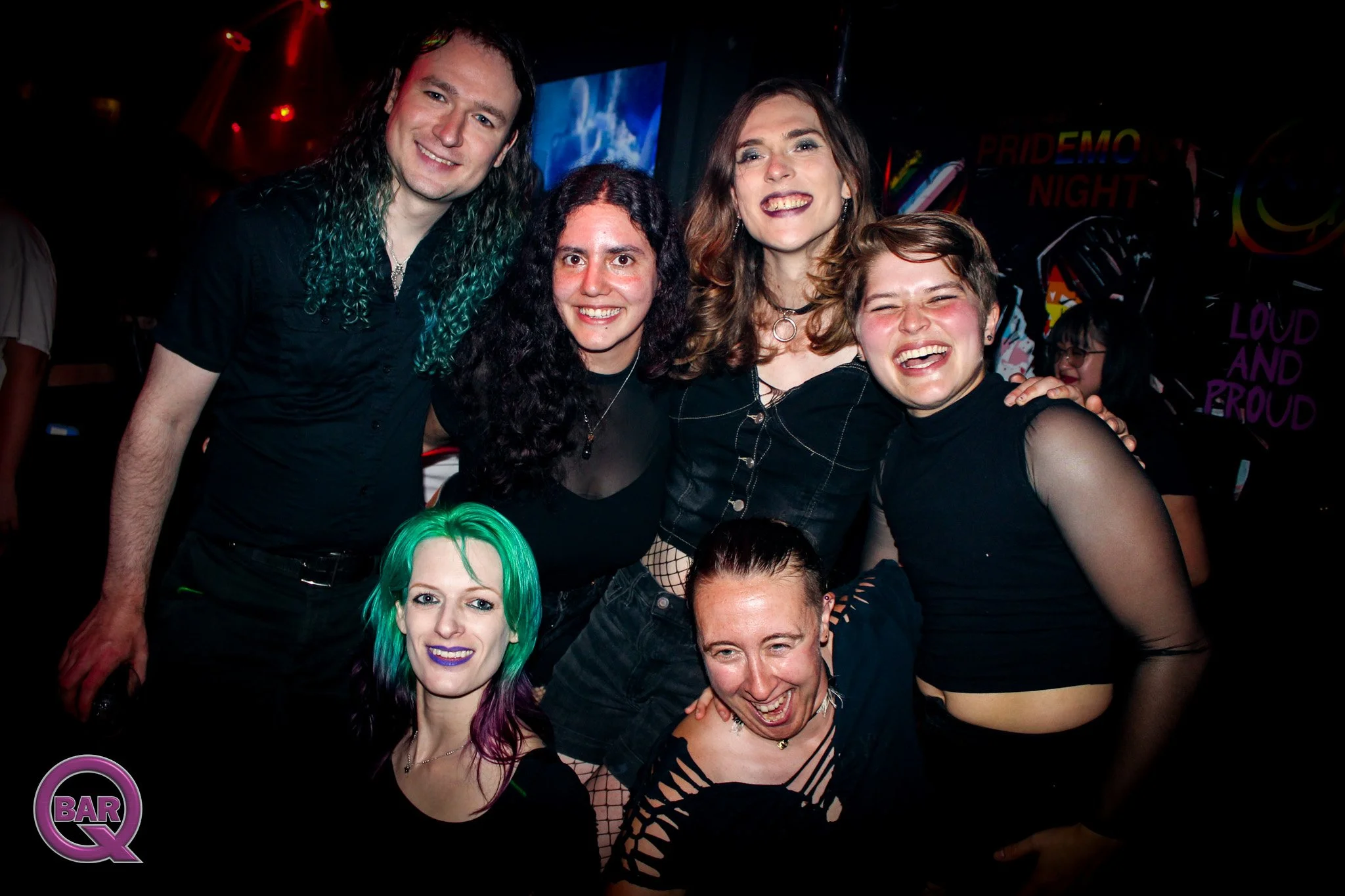 Group of people enjoying a night out at a bar or club, smiling and posing for the camera, with colorful lighting and a sign that reads 'PRIDE DEMO NIGHT' in the background.