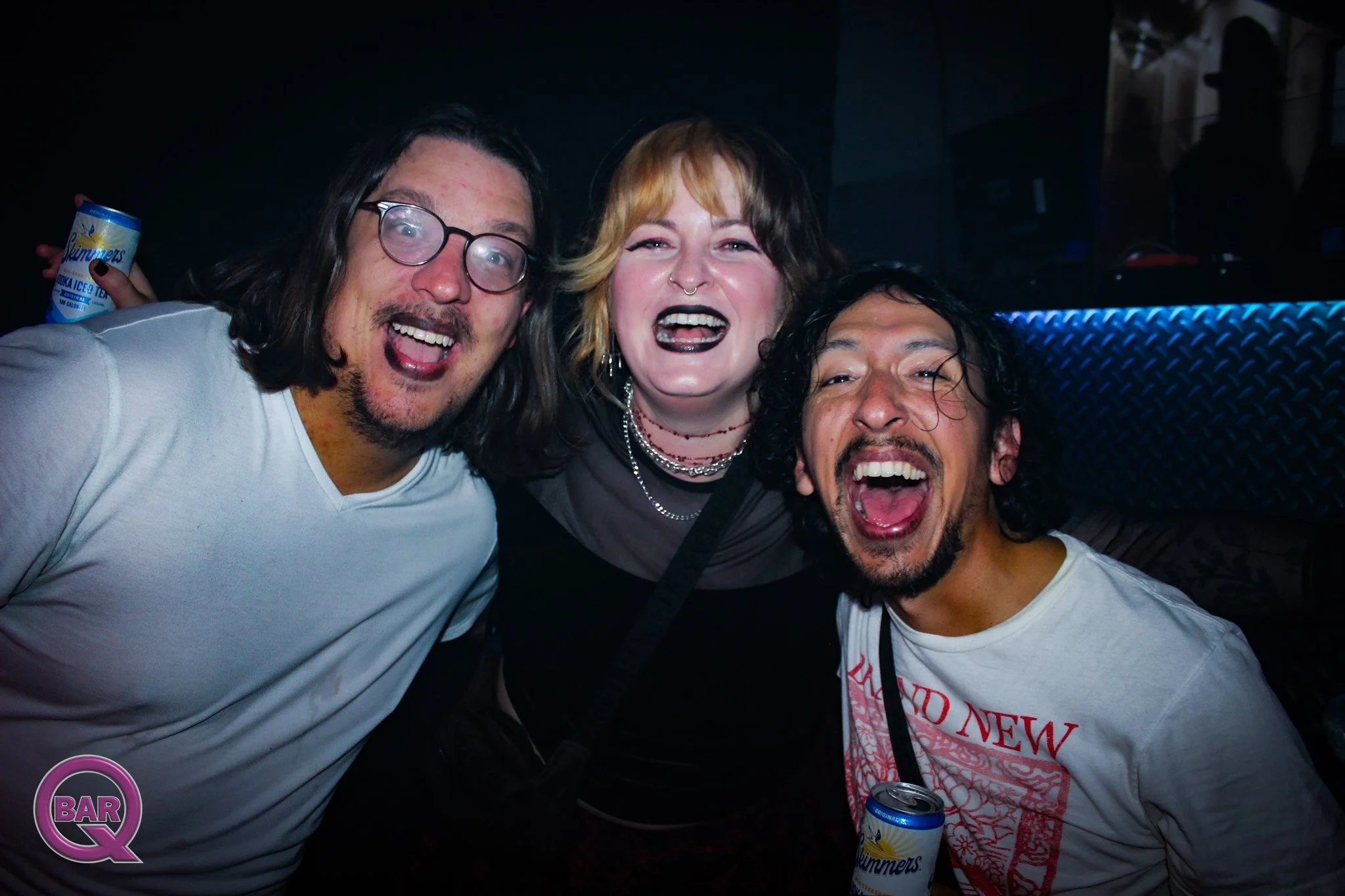 Three friends happily taking a selfie together at a bar or nightclub, with dark background and a visible logo that says 'BAR Q' in the bottom left corner.