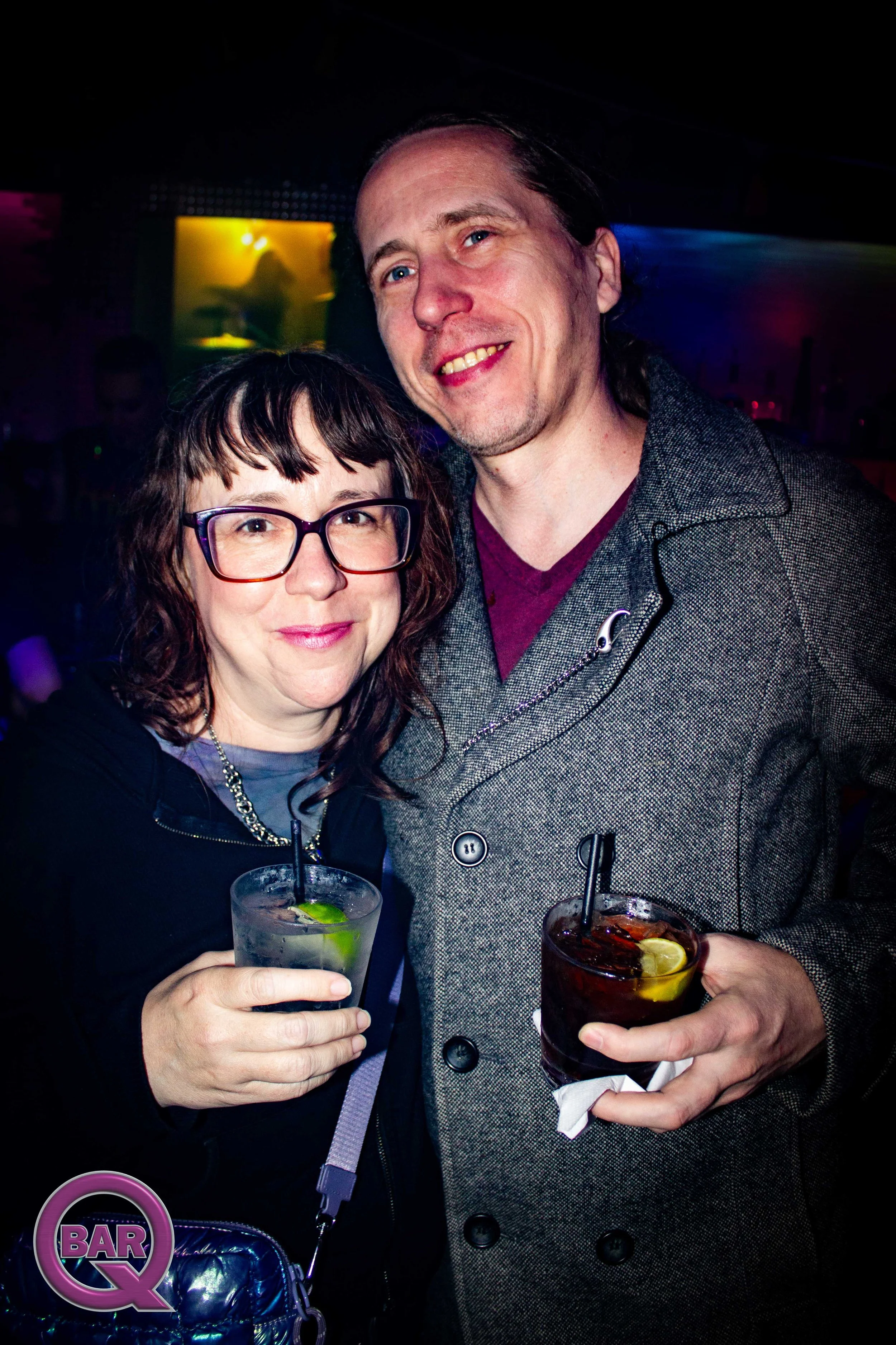 A woman and a man smiling and holding drinks in a bar or nightclub with colorful lighting in the background.