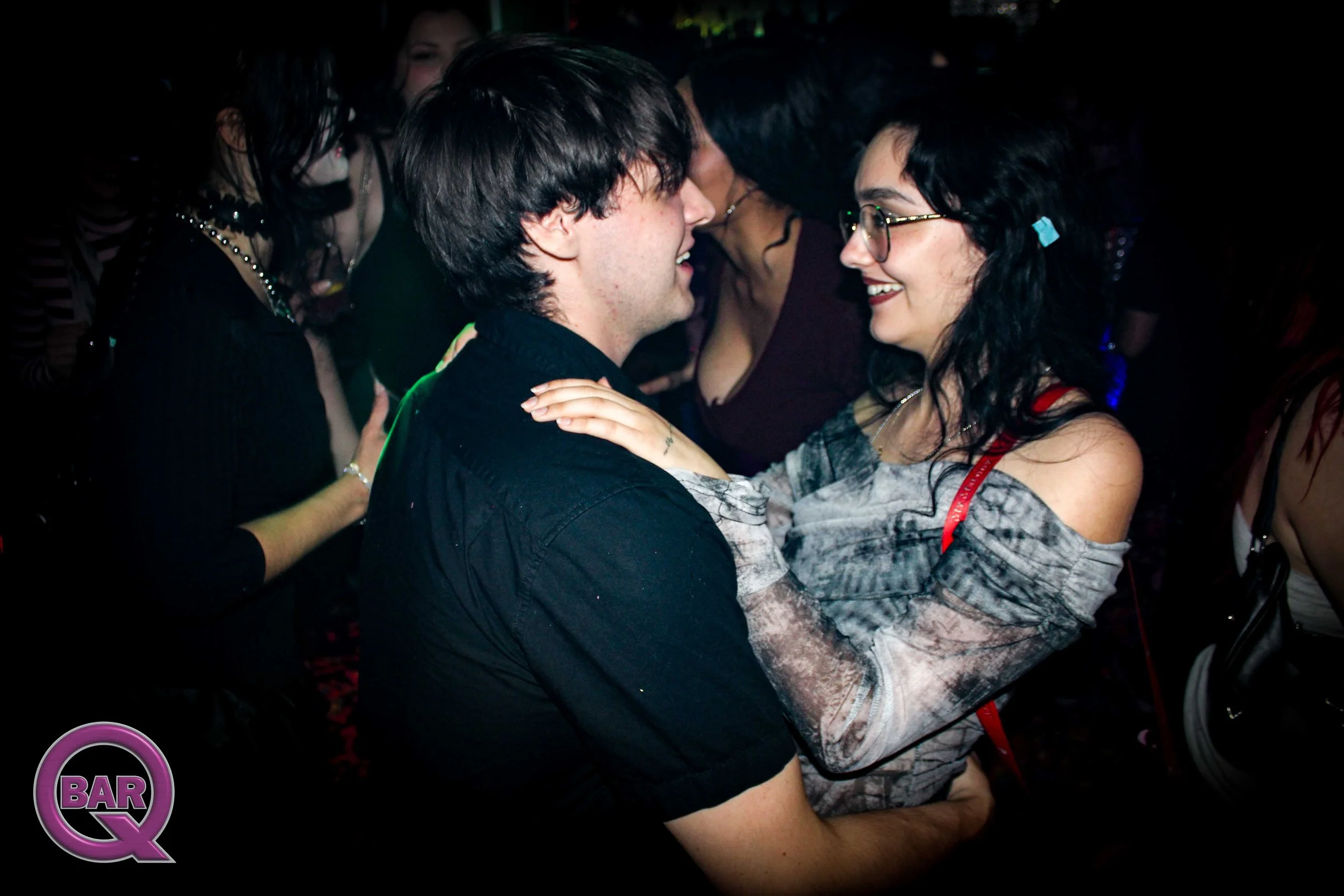 A young couple dancing closely at a nightclub, surrounded by other people, with a dark background and colorful lights.