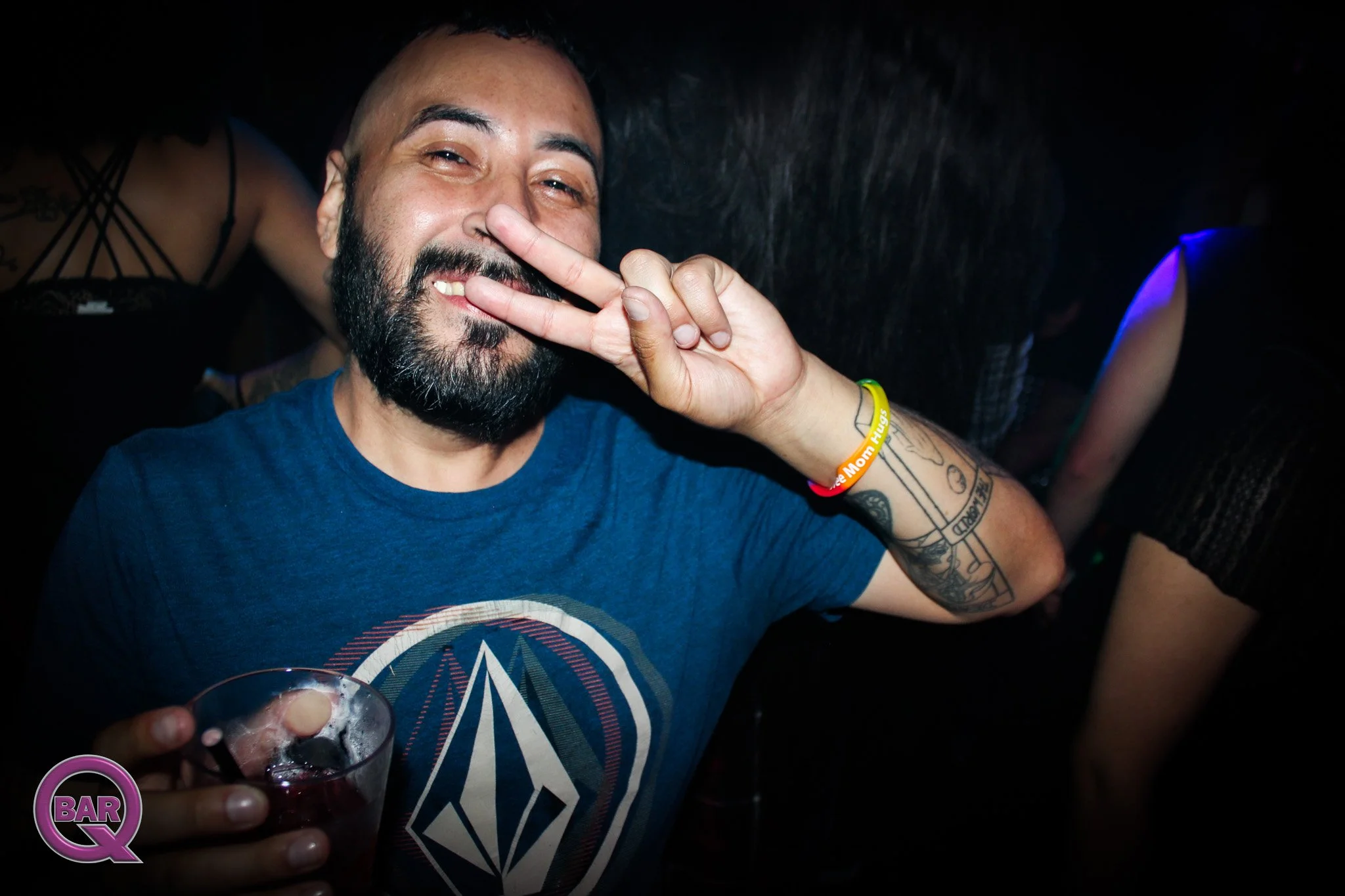 A man with a beard and tattoos smiling and making a peace sign near his face. He is holding a glass of dark-colored drink with ice and a straw. The background is dark and blurred, indicating a bar or club setting.