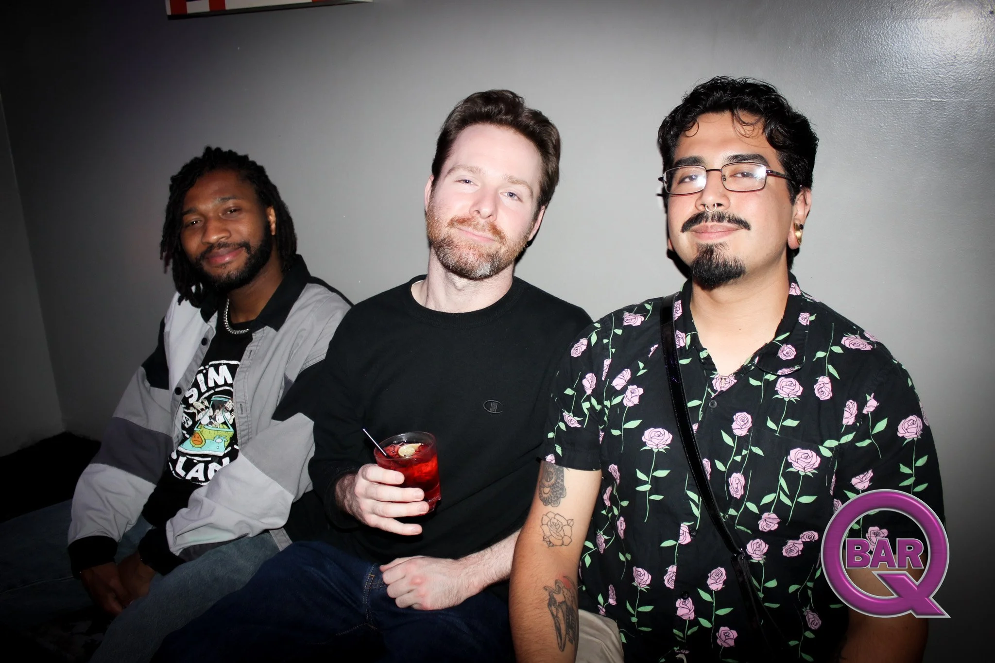 Three men sitting at a bar with a dark gray wall background. The man in the middle is holding a red cocktail and smiling, wearing a black T-shirt. The man to the right wears glasses and has tattoos on his arms, wearing a black shirt with pink roses. 