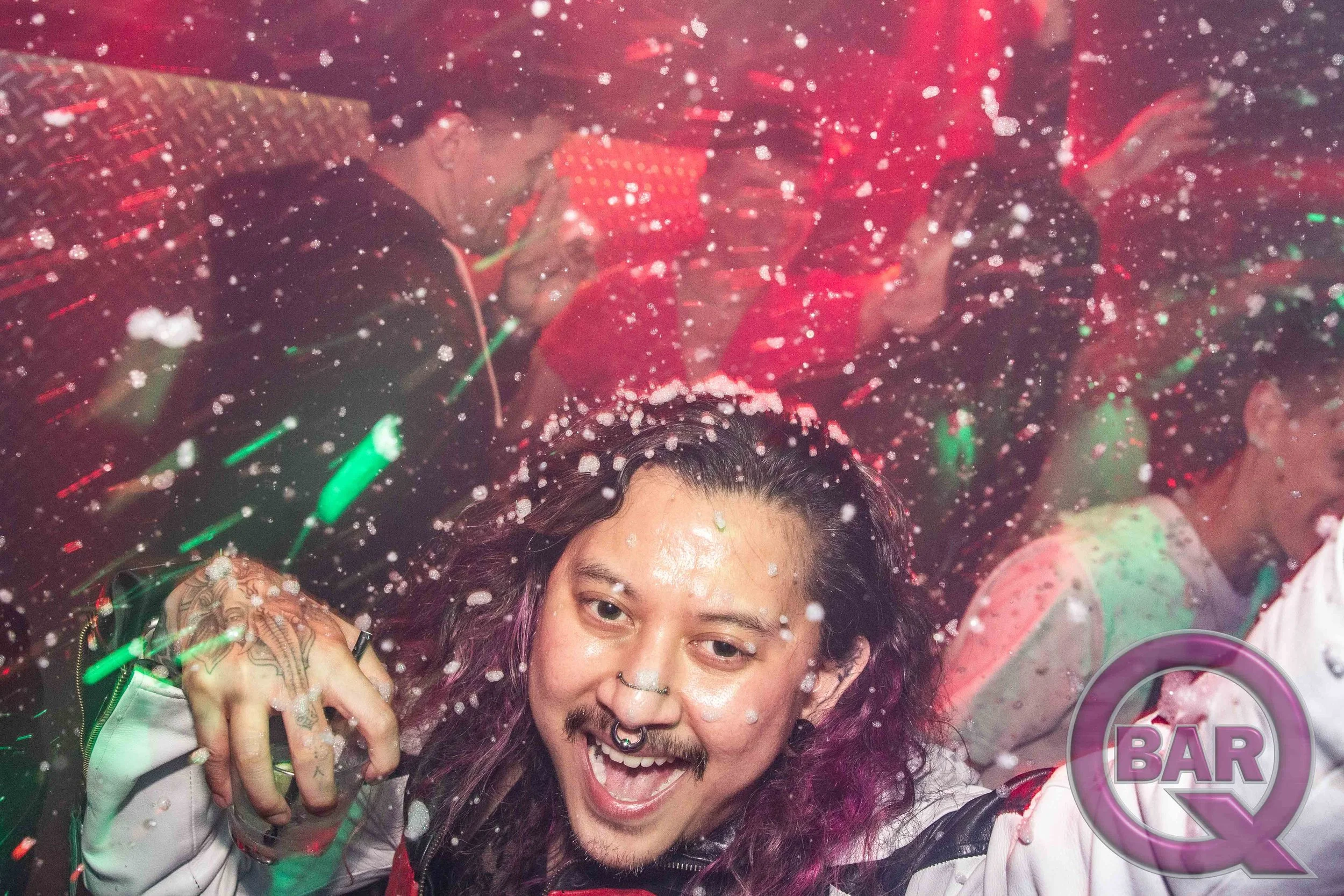 A person with long dark hair and multiple facial piercings smiling, surrounded by colorful party decorations and confetti.