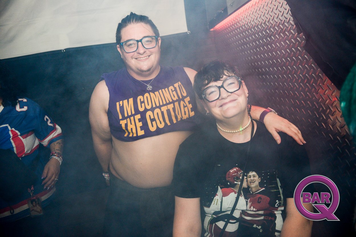 Two smiling individuals at a bar or club, one wearing a sleeveless shirt that says "I'm Coming to the Cottage," and the other wearing glasses and a hockey jersey with sports posters on it, with a "BarQ" logo in the bottom right corner.