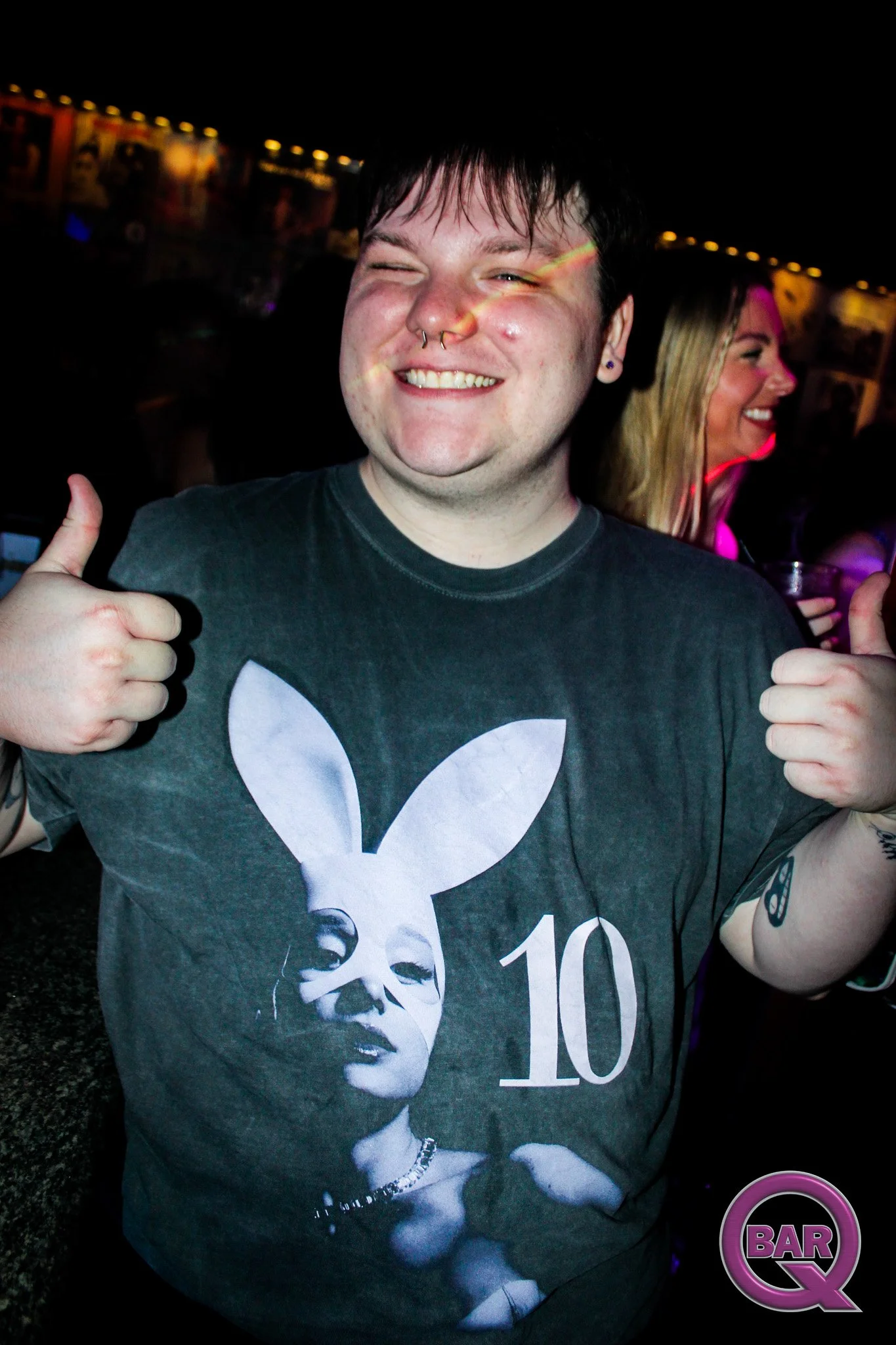 A young man smiling and giving two thumbs up. He has dark hair, a septum piercing, and is wearing a black t-shirt with a stylized black and white portrait of Marilyn Monroe and the text '10'. In the background, a woman with blonde hair is also smilin