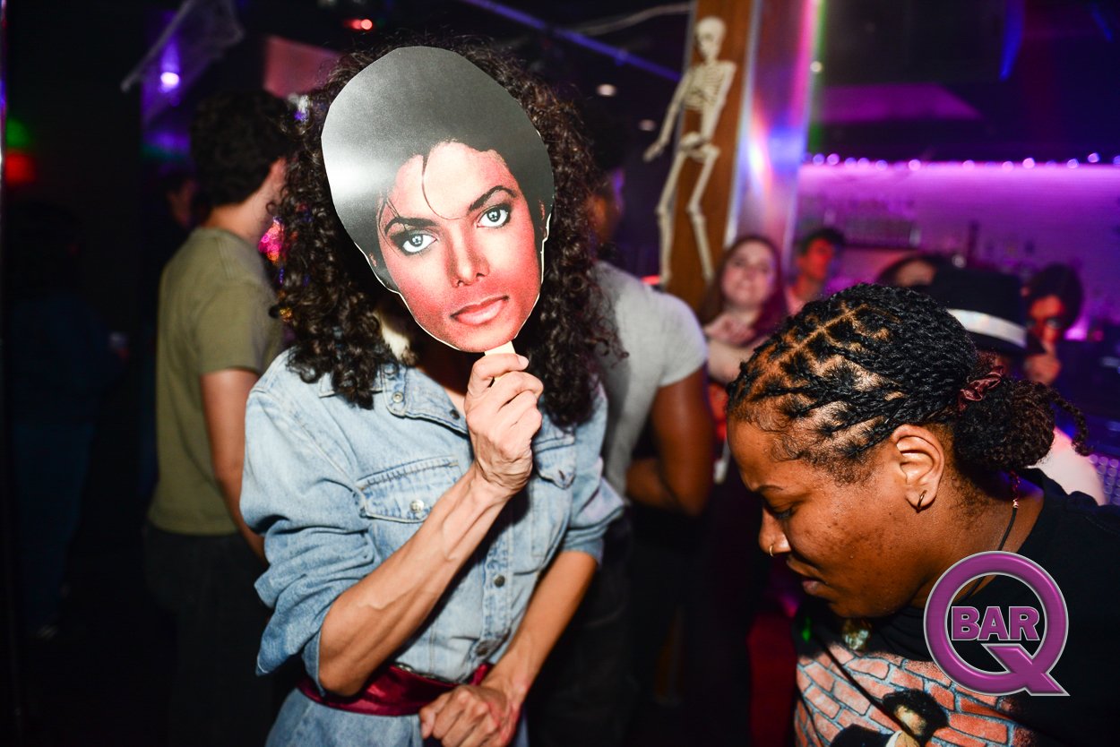 Person holding a large mask of a woman's face over their own face at a party or nightclub with colorful lighting and other people in the background.