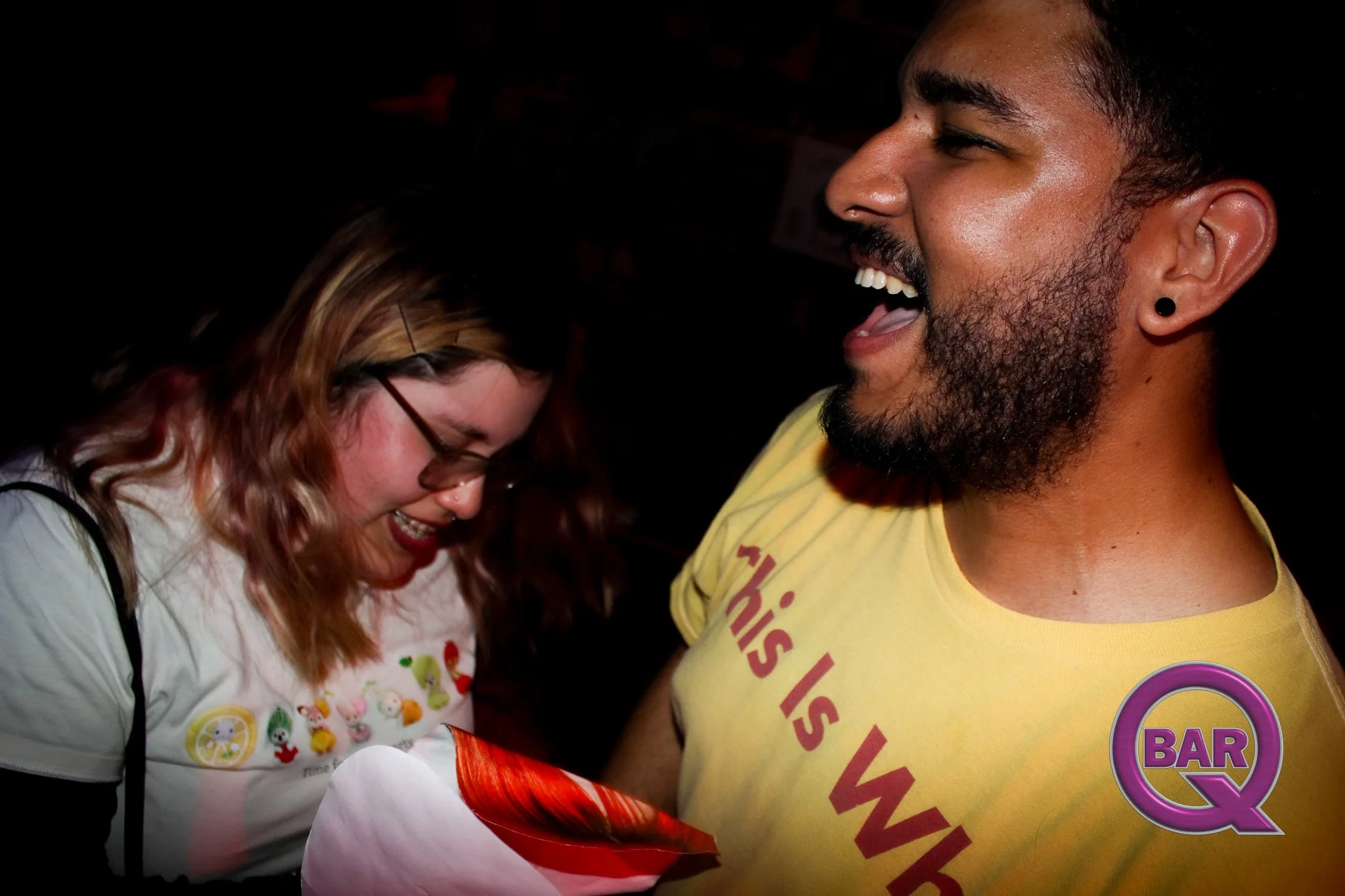 A man and woman sharing a moment of laughter, the woman is wearing glasses and a white shirt with colorful characters, and the man is wearing a yellow shirt with text and a purple badge.