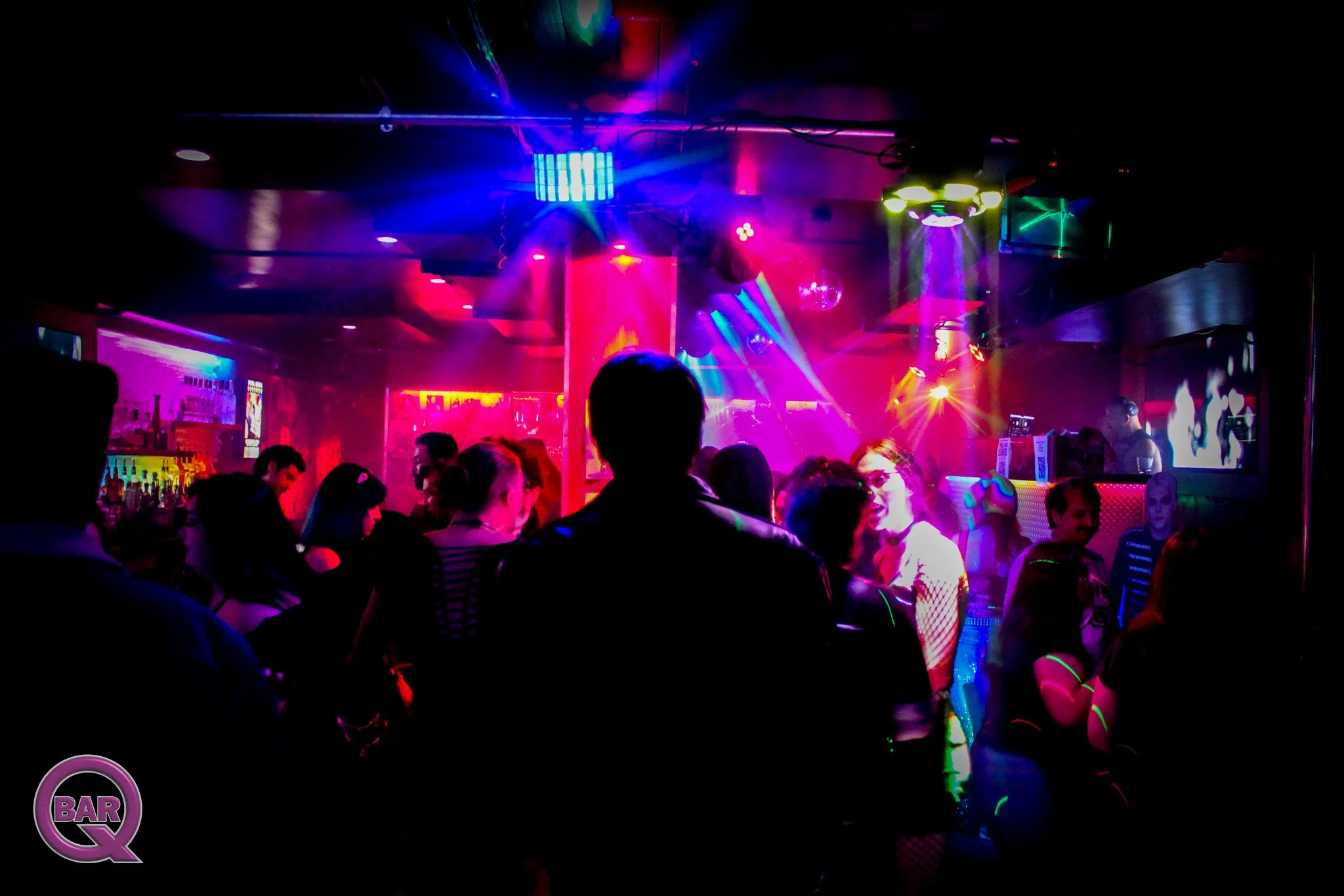People dancing and socializing in a nightclub filled with colorful lights and a bar area.