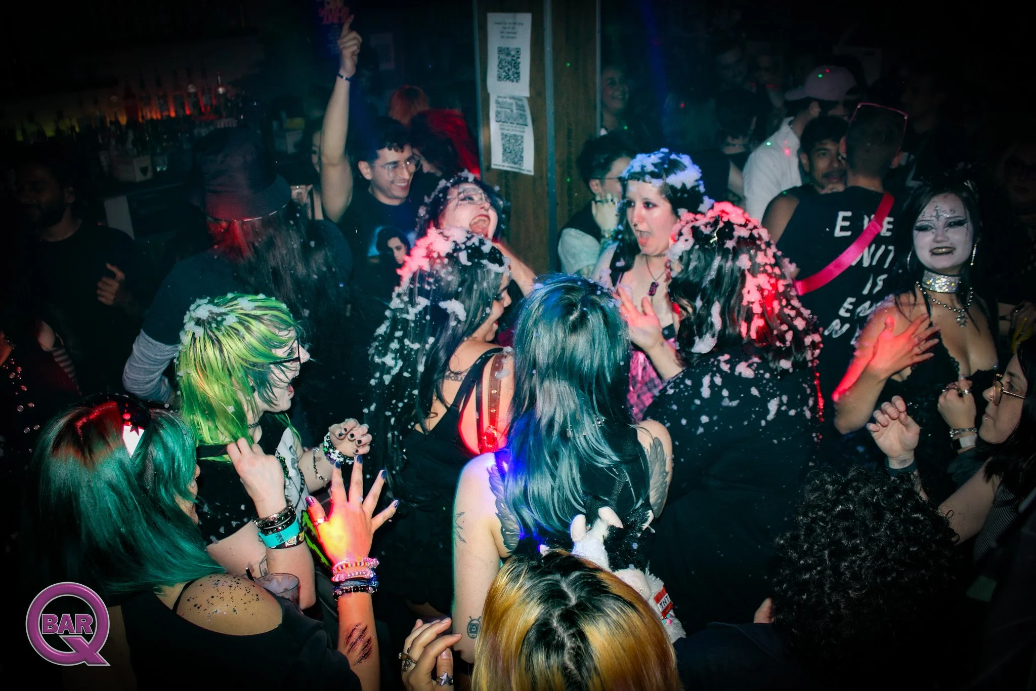 People dancing and celebrating at a crowded party or nightclub, with some wearing colorful wigs and makeup, and foam or bubbles in the air.