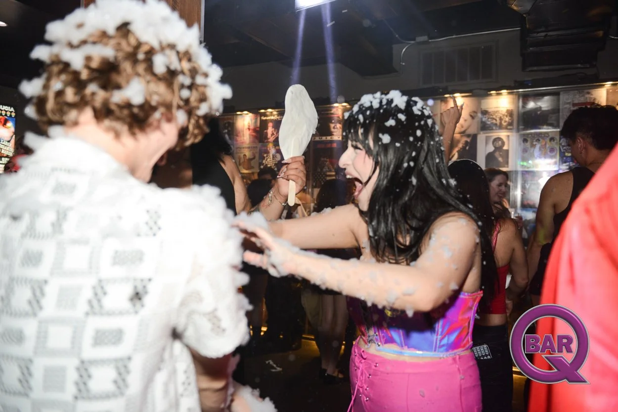 Two women are celebrating at a party, with one woman holding a popsicle and the other throwing foam or confetti on the woman with the popsicle, inside a lively bar or club with posters on the wall and other people in the background.