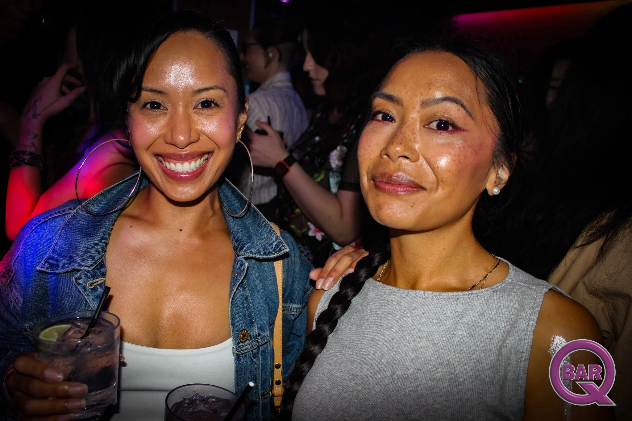 Two women smiling and close together at a social event, with a dark background and other people in the distance.