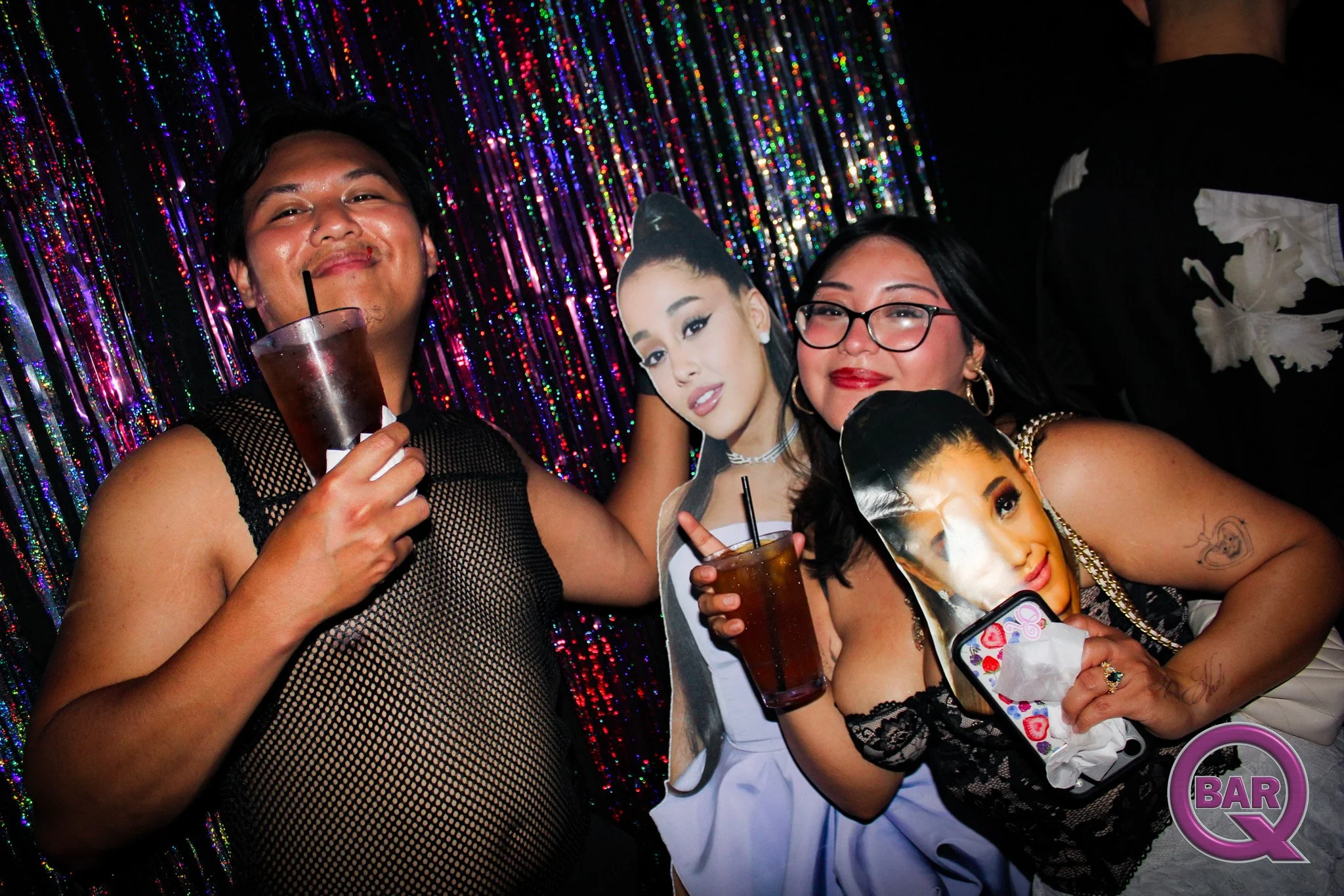 Group of people celebrating at a party with colorful lights and holding drinks, with a backdrop of shiny, multicolored, vertical strands.