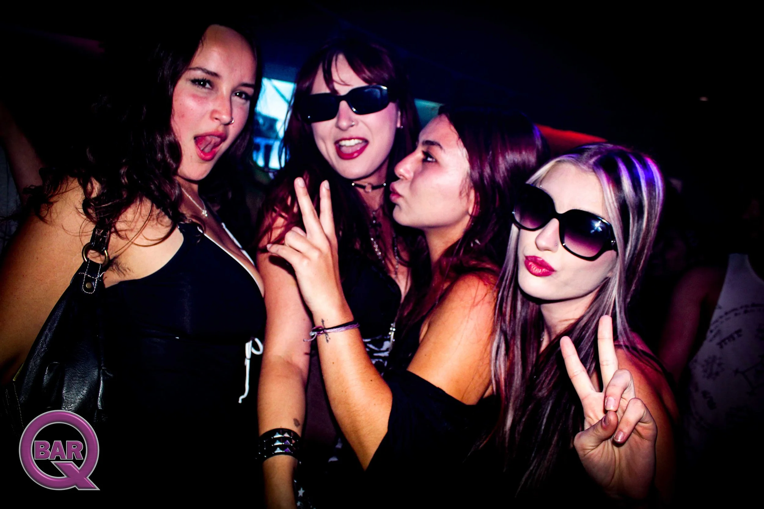 Four women posing closely together in a dimly lit nightclub, some wearing sunglasses and making peace signs.