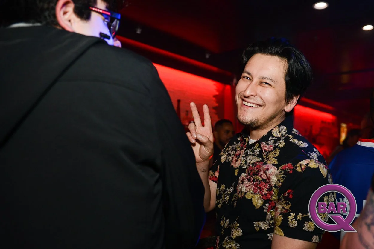 A man smiling and making a peace sign with his hand at a bar or nightclub, with a red-lit background and a purple and pink 'Bar Q' logo in the bottom right corner.