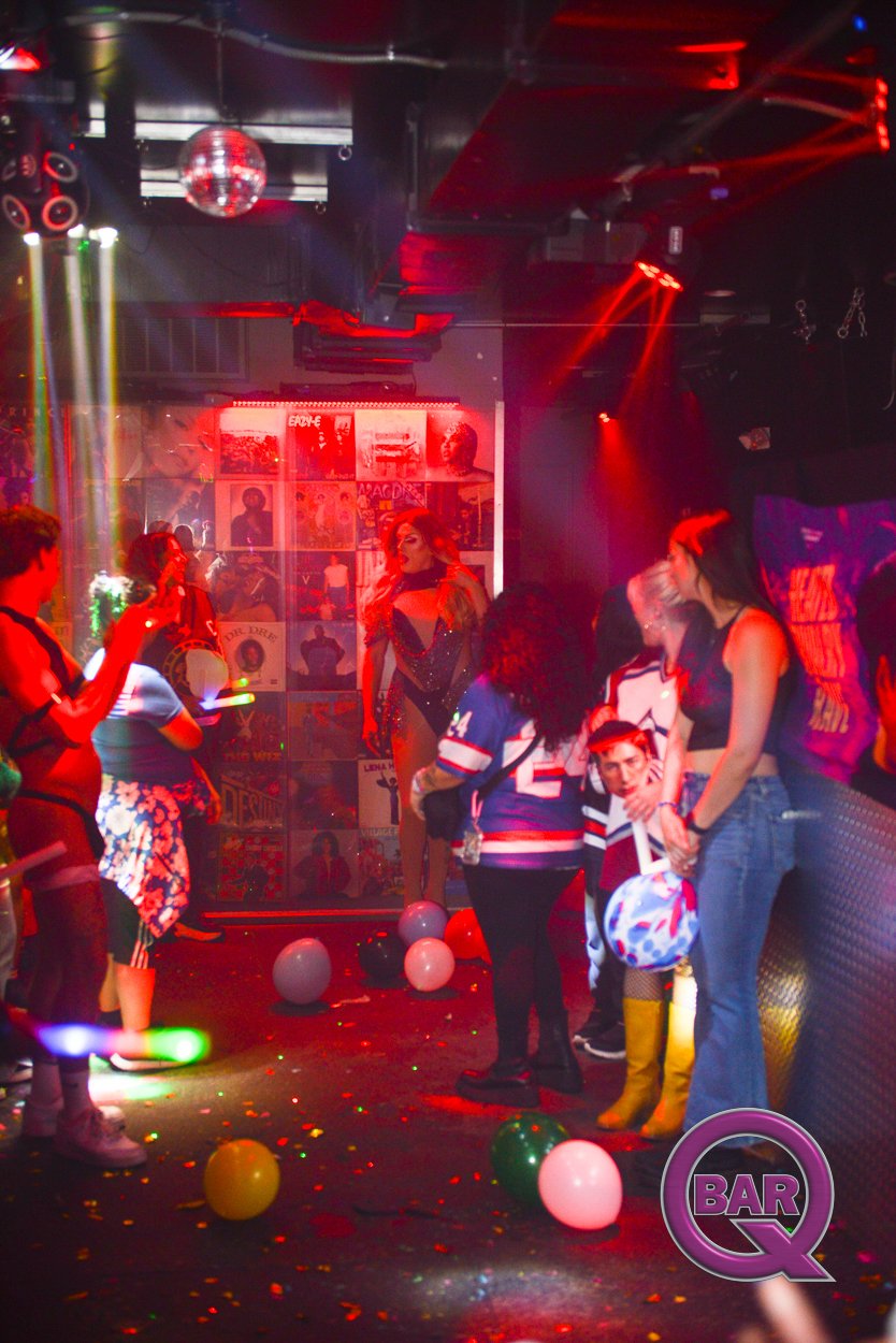 People dancing and socializing in a dimly lit nightclub with colorful lights, balloons, and posters on the wall.