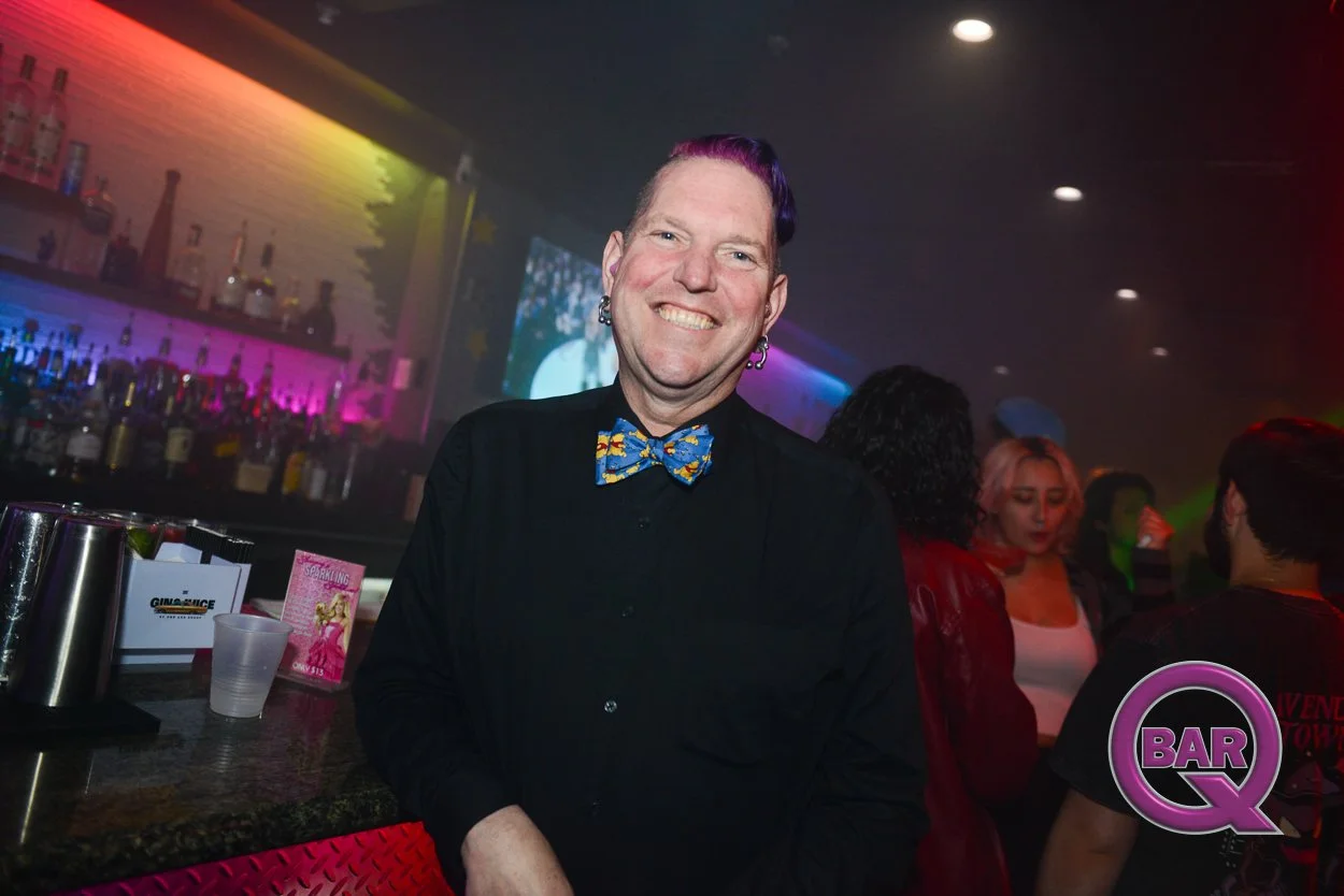 A smiling man with purple hair and earrings, wearing a black shirt and colorful bowtie, standing at a bar in a nightclub with neon lighting and other people socializing in the background.