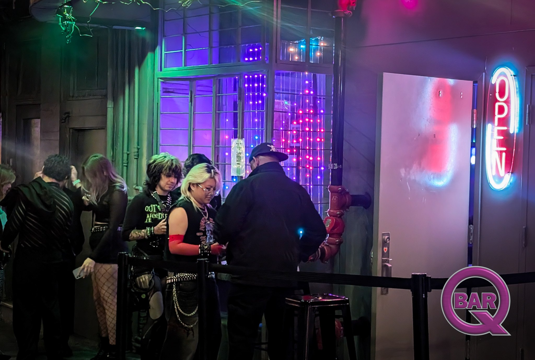 A group of people standing outside a nightclub or bar with neon signs and colorful lights, waiting in line or in front of the entrance.