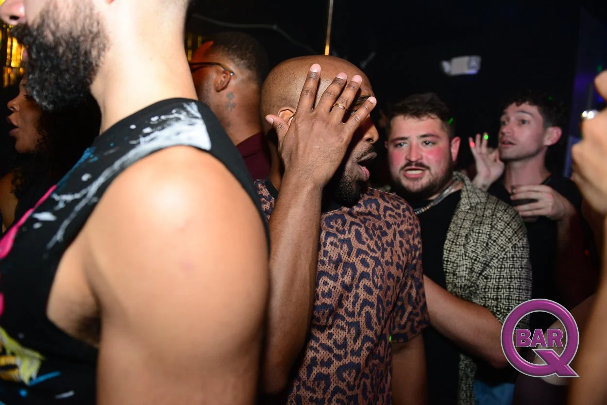 Group of people dancing and socializing at a nightclub or party, with some making expressive gestures and faces, illuminated by colorful lights, with a 'Bar Q' logo in the bottom right corner.