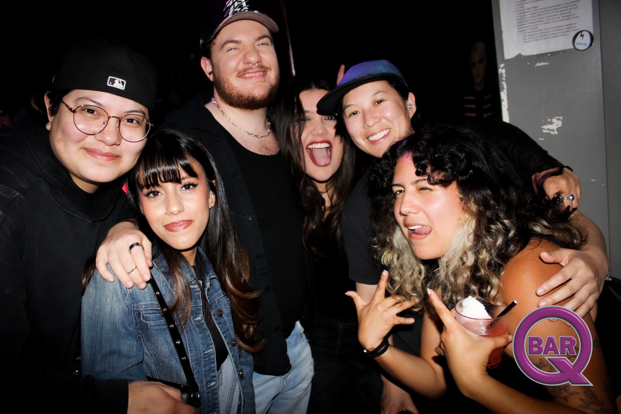Group of six young adults smiling and posing for a photo at a bar or nightclub, with some making playful gestures.