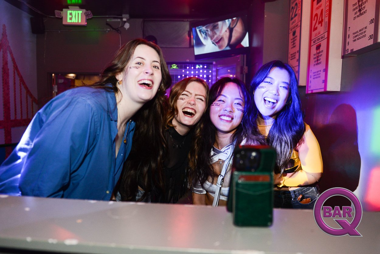 Four women taking a selfie with a smartphone in a bar, smiling and laughing. The bar has colorful neon lights, an exit sign, and menu boards visible in the background.