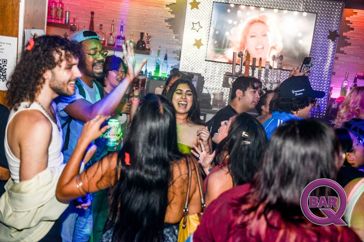 Group of young people dancing and partying at a bar or nightclub with colorful lighting and a large screen displaying a woman's face in the background.