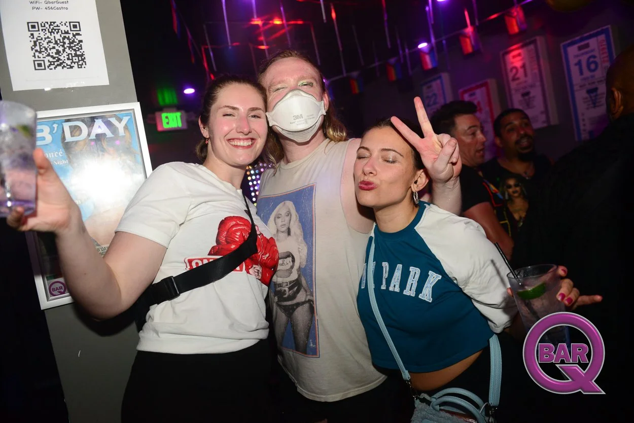 Three young women celebrating at a bar, smiling and making playful gestures, with disco lights and bar-themed posters in the background.
