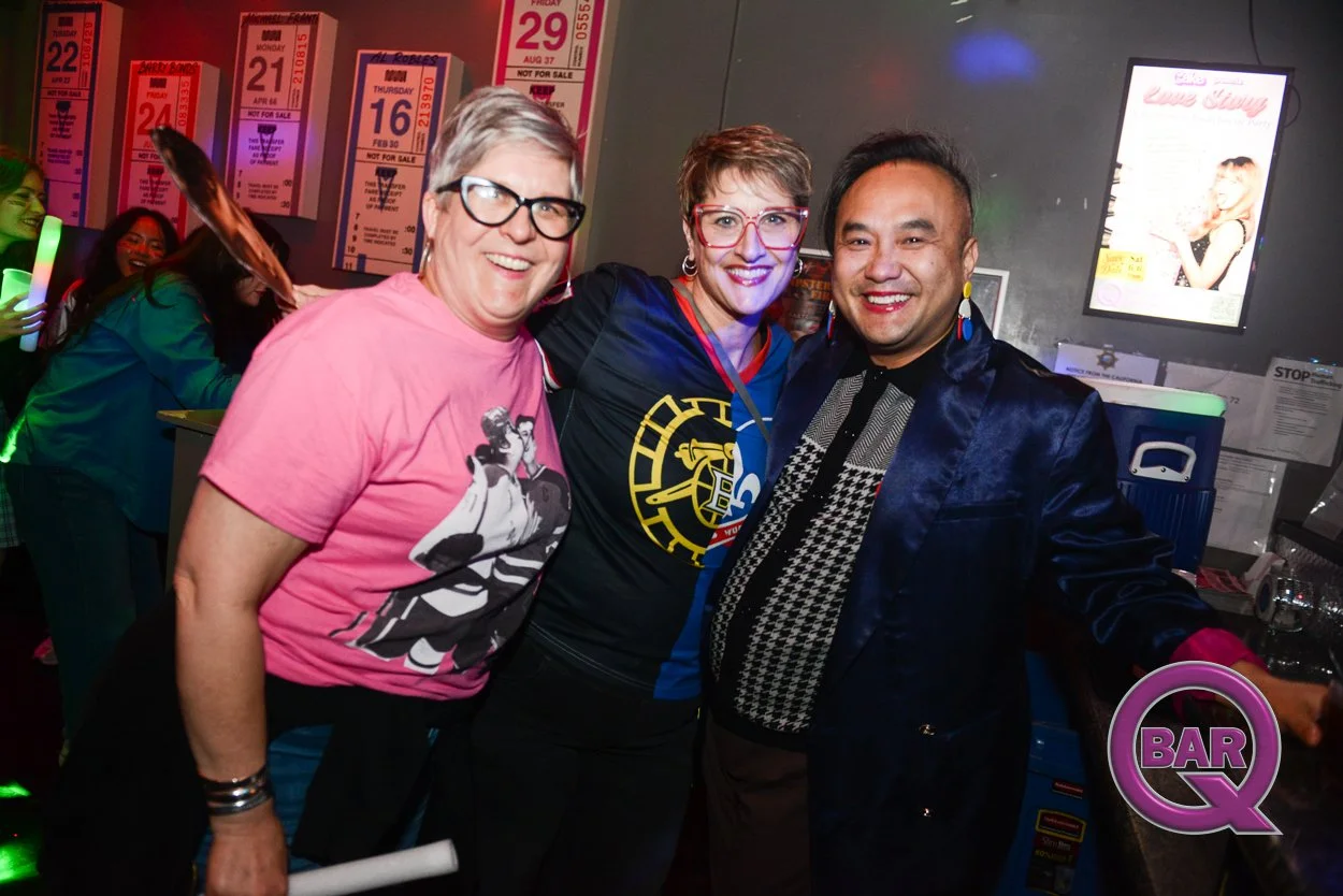 Three people smiling and posing for a photo at a bar, with a group of people in the background, bar signs, and a television screen.