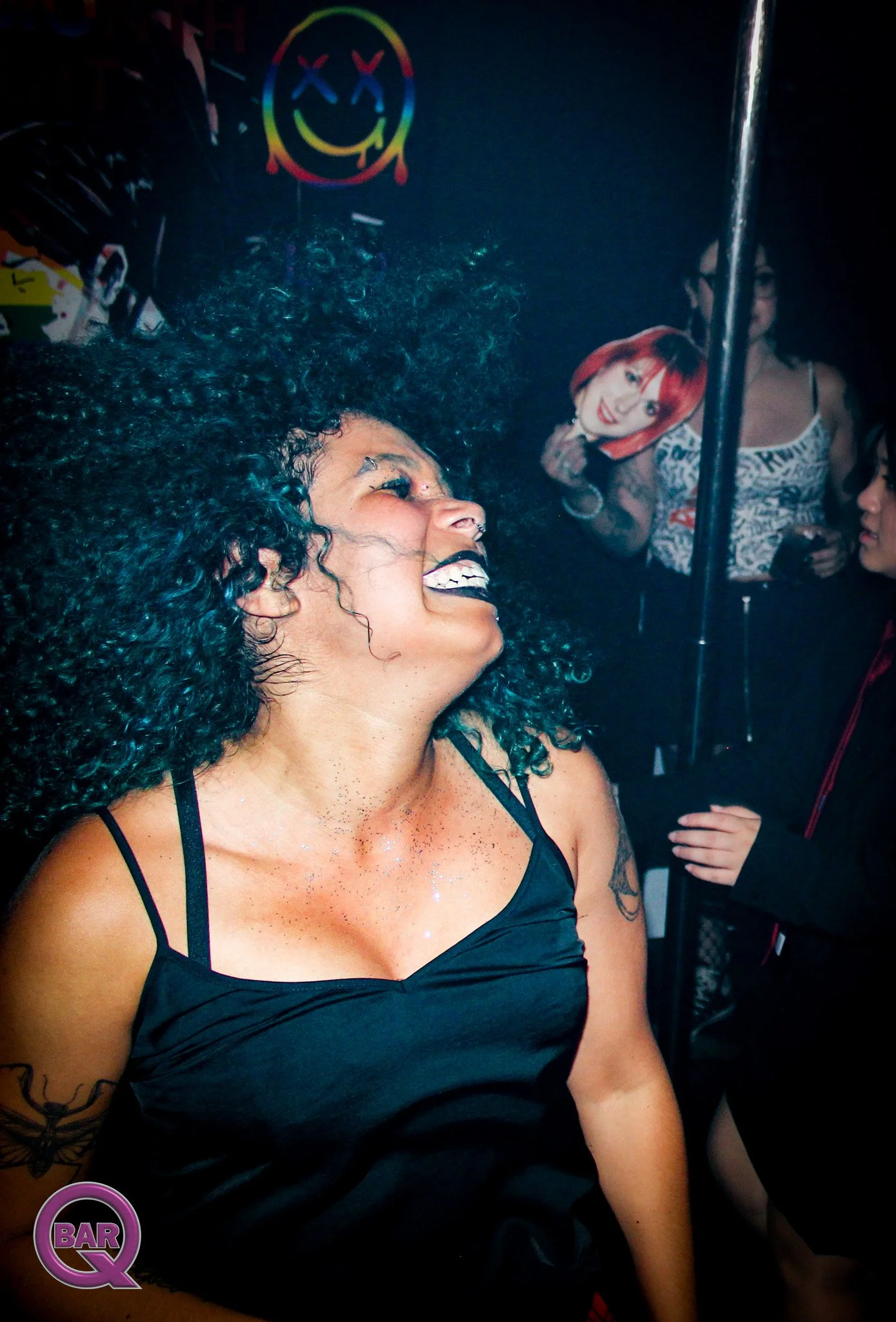 A woman with curly black hair and a black top smiling, with multiple women in the background, some holding drinks, inside a bar or club with neon signs.
