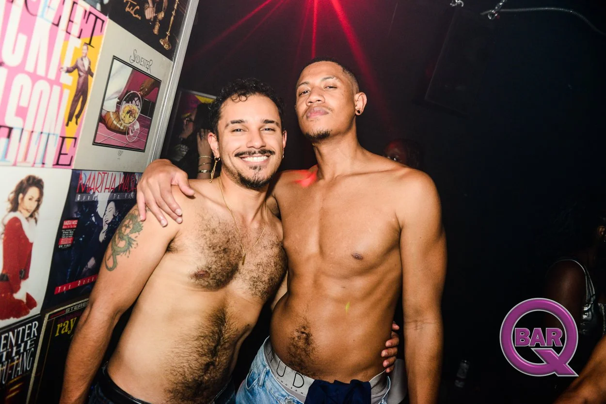 Two shirtless men smiling and embracing in a nightclub, with posters on the wall and a dark background.