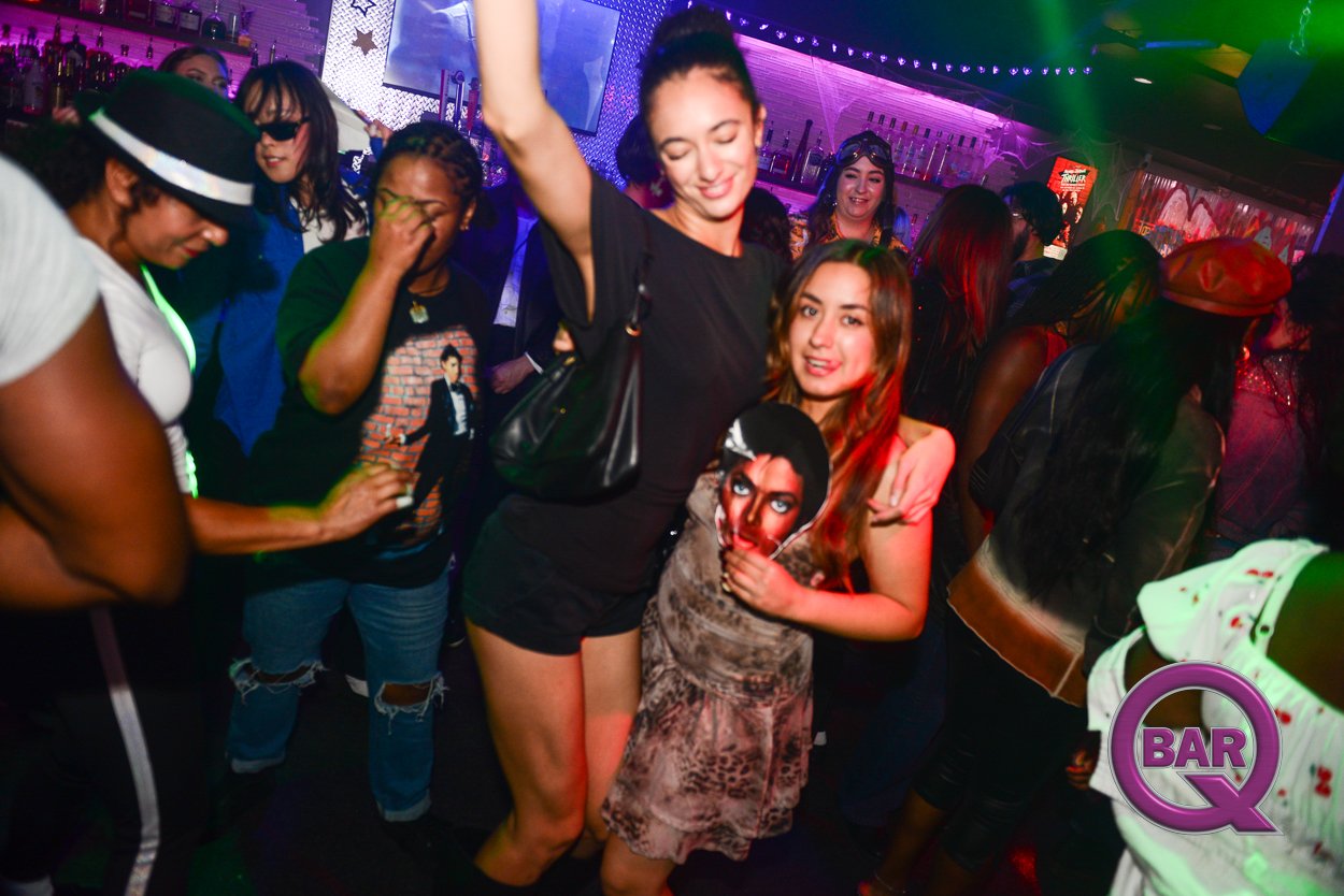 Group of diverse women dancing and enjoying themselves at a nightclub with colorful lighting, bar shelves, and a lively atmosphere.