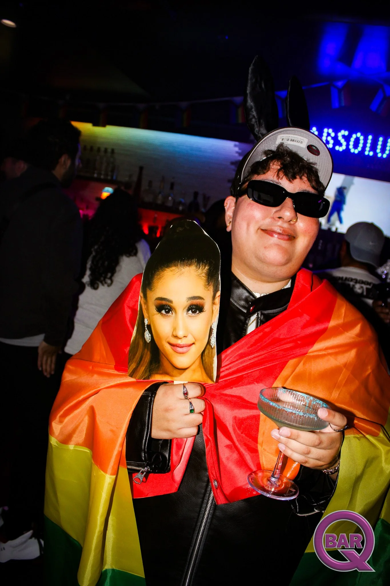 A person with short curly hair wearing dark sunglasses, a nose ring, and a rainbow flag cape at a bar, holding a cardboard cutout of a woman's face and a glass of drink in a dimly lit venue.
