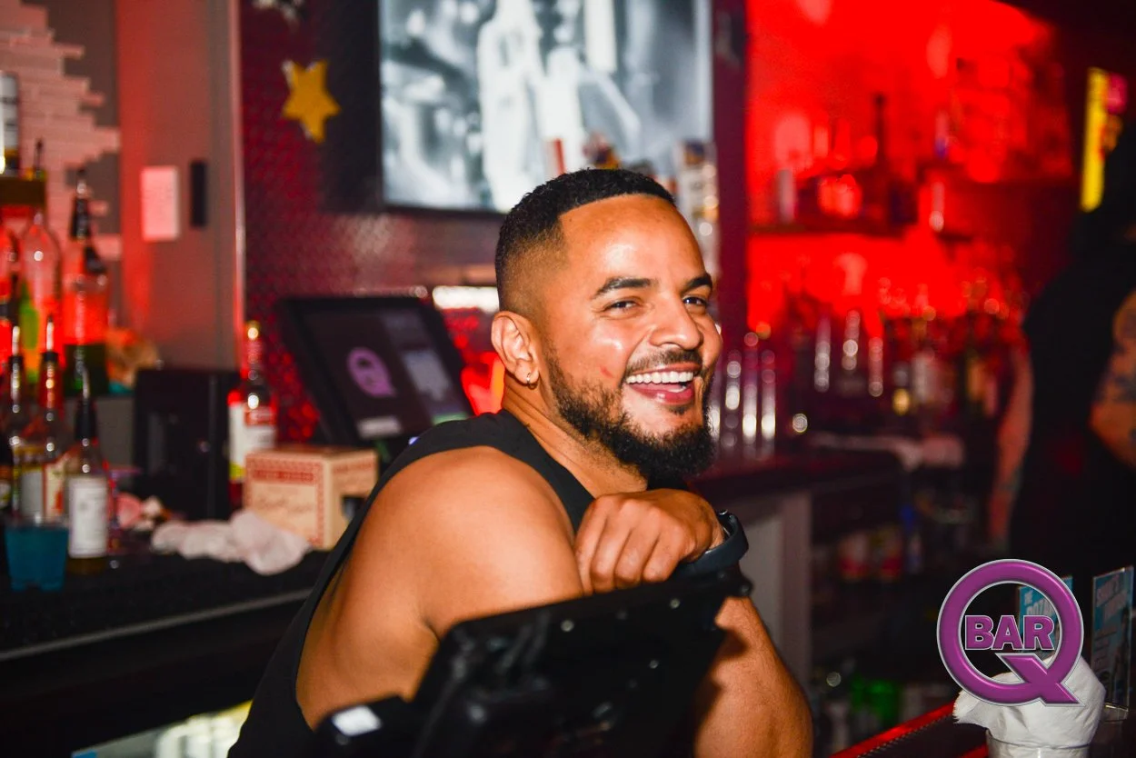 A smiling man with a beard and short hair at a bar, with bottles and bar equipment in the background, illuminated by red and purple lighting.