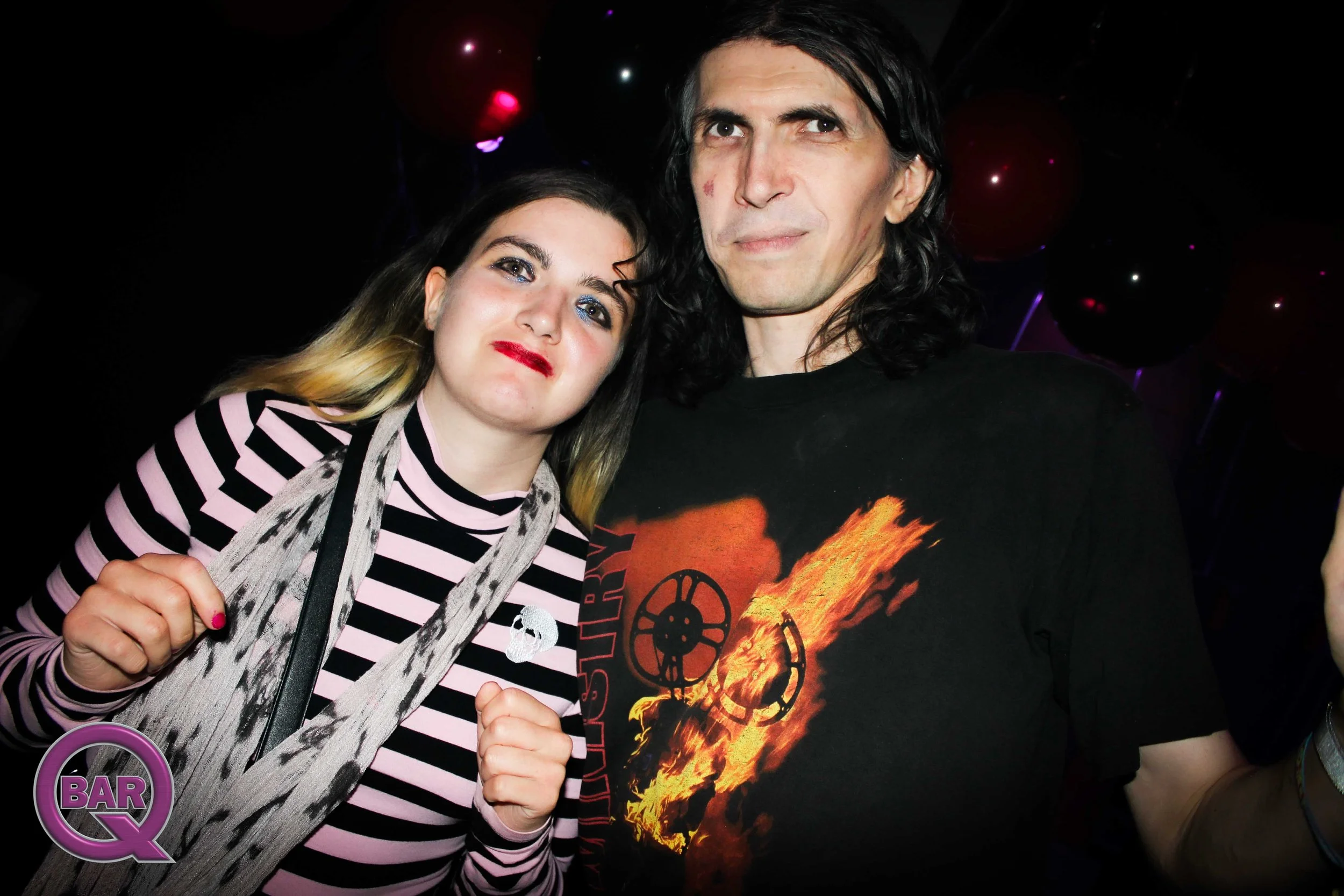 A young woman with makeup and dreadlocks poses next to a man wearing a graphic t-shirt with flames and a skeleton motorcycle. The woman is holding a drink, and both appear to be at a party or bar, with balloons and a dark background.