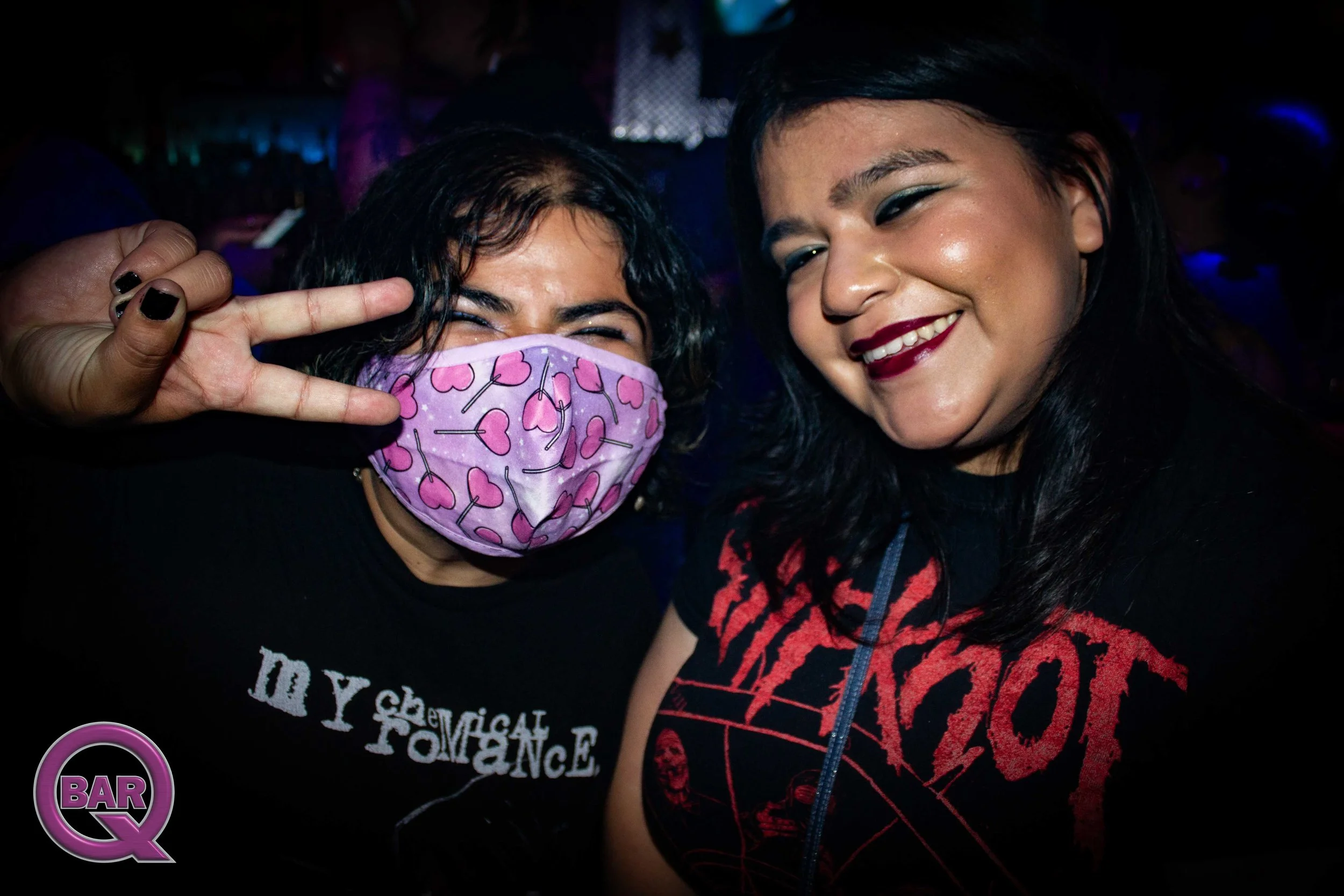 Two women at a bar, one wearing a colorful face mask with hearts and the other with dark lipstick, both smiling and posing for a photo.