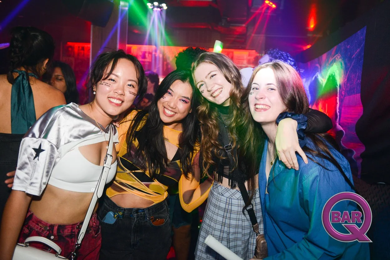 Four young women smiling and posing together at a nightclub, with colorful lights and a vibrant atmosphere.