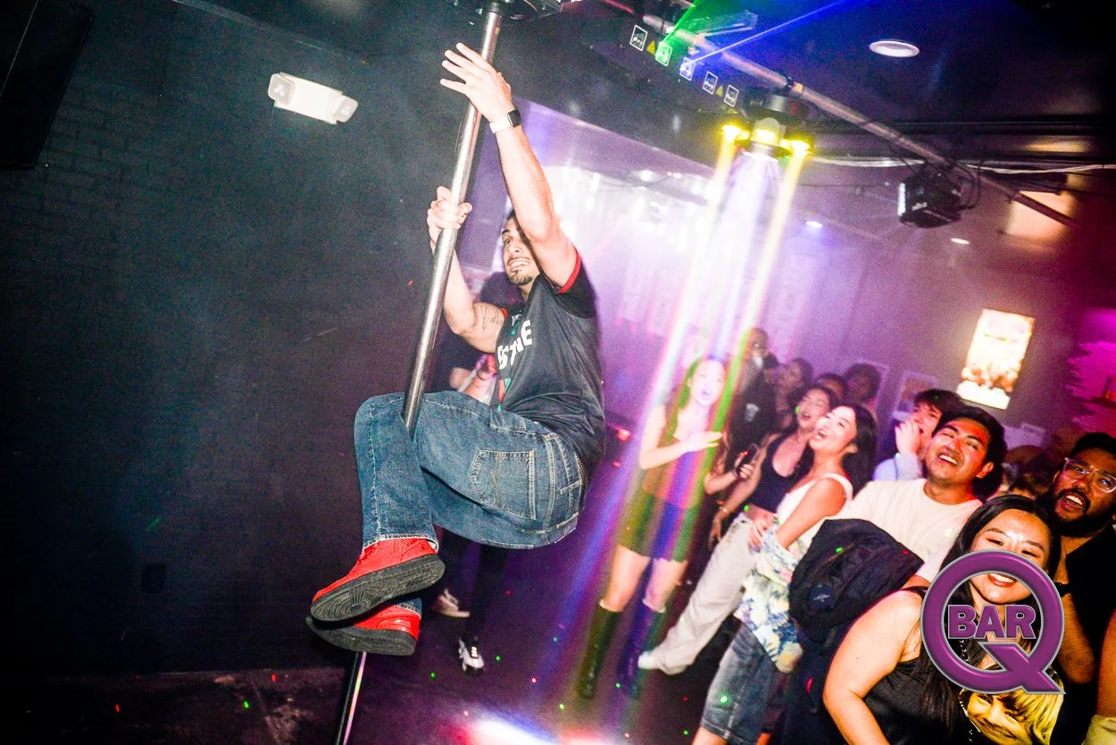 A man riding a pole in a nightclub with a crowd watching and smiling in the background, colorful lighting overhead.