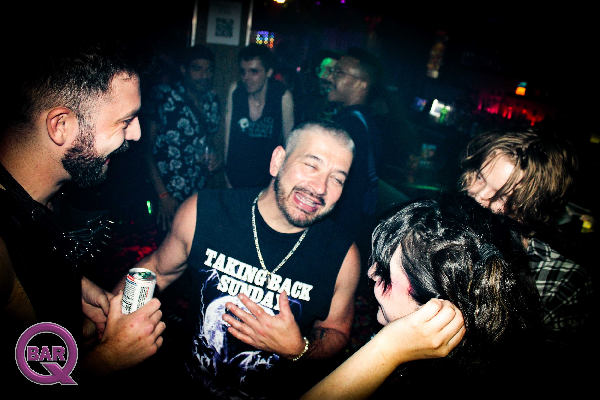 Group of people smiling and dancing in a nightclub with colorful lights, some holding drinks, and a bar visible in the background.