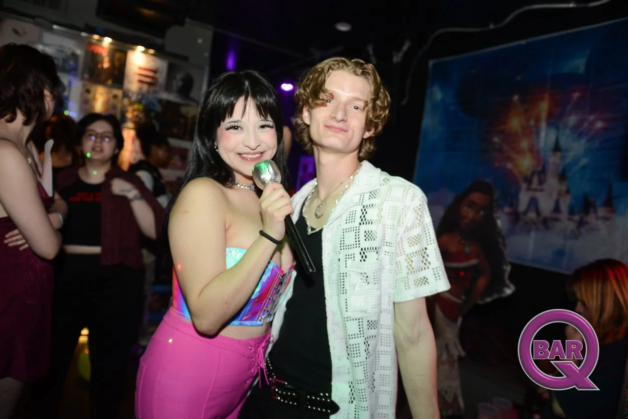 A group of people at a bar or nightclub, with two individuals in the foreground posing for a picture. The woman on the left is smiling and holding a microphone, wearing a colorful strapless top and pink pants. The man on the right has curly hair, is 