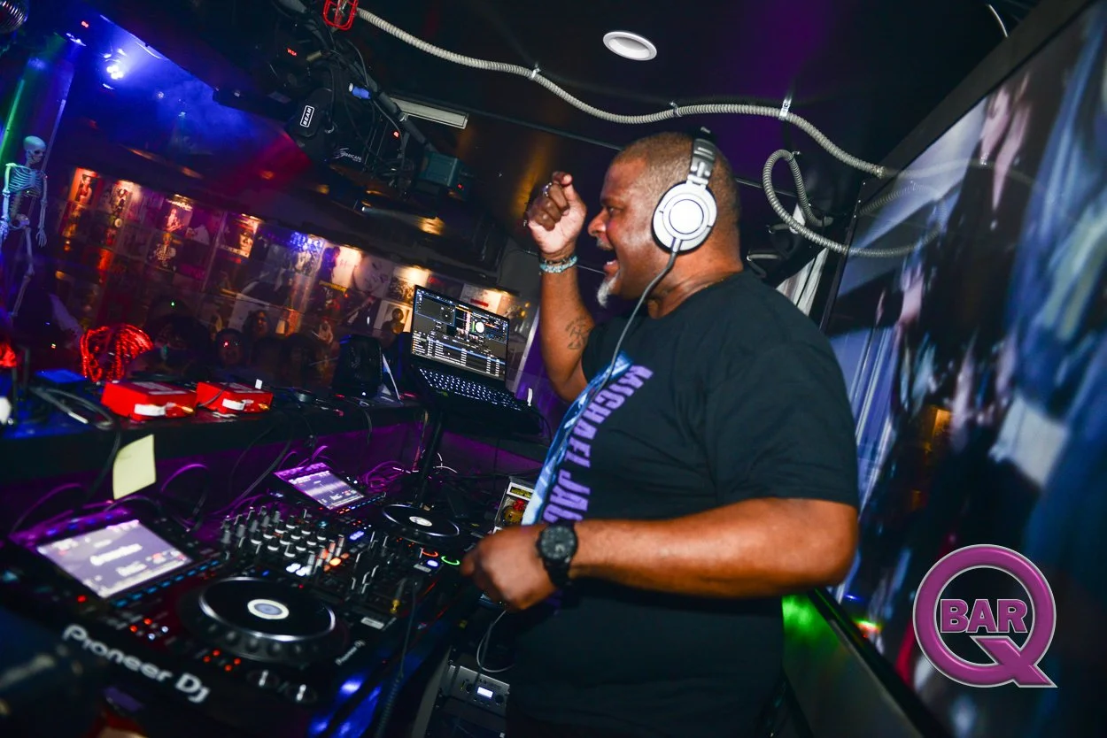 A DJ wearing headphones and a black t-shirt is playing music at a nightclub, with a crowd dancing in the background and colorful lights illuminating the scene.