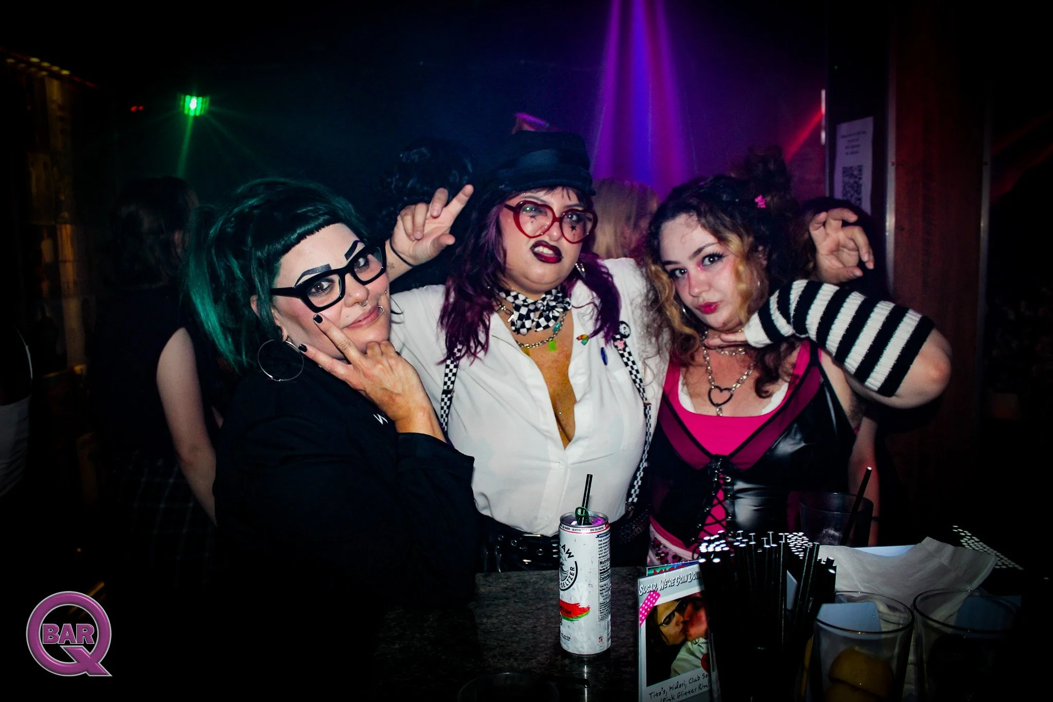 Three women dressed in colorful and alternative fashion pose together at a nightclub or party, with vibrant lights and a DJ setup in the background.