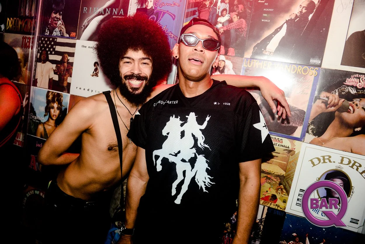 Two men at a music or record store, standing in front of a wall filled with various vinyl album covers, one shirtless with big afro, the other wearing sunglasses and a black graphic T-shirt.