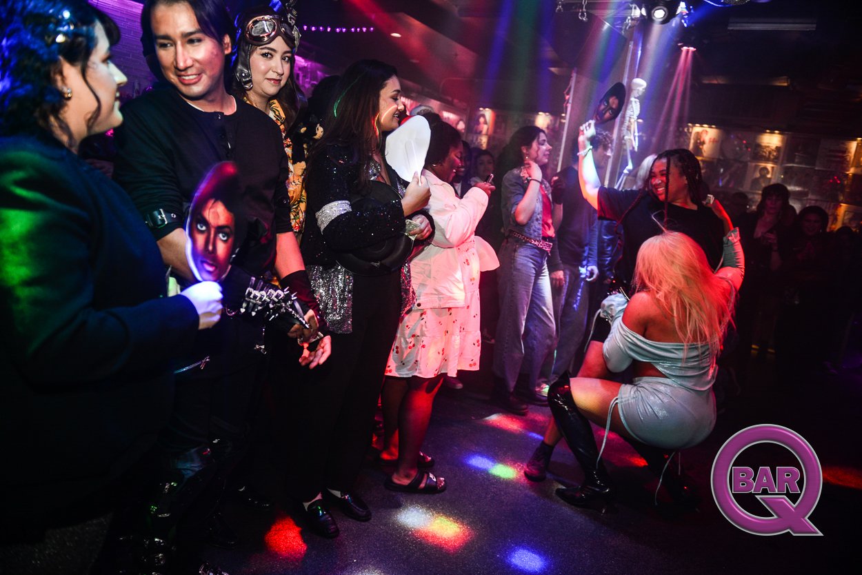 People dancing and socializing in a nightclub with colorful lights and decorations, including a skeleton figure on the stage. One woman is kneeling and dancing with a big smile, while others chat and watch the performance. There is a "Bar Q" logo in 