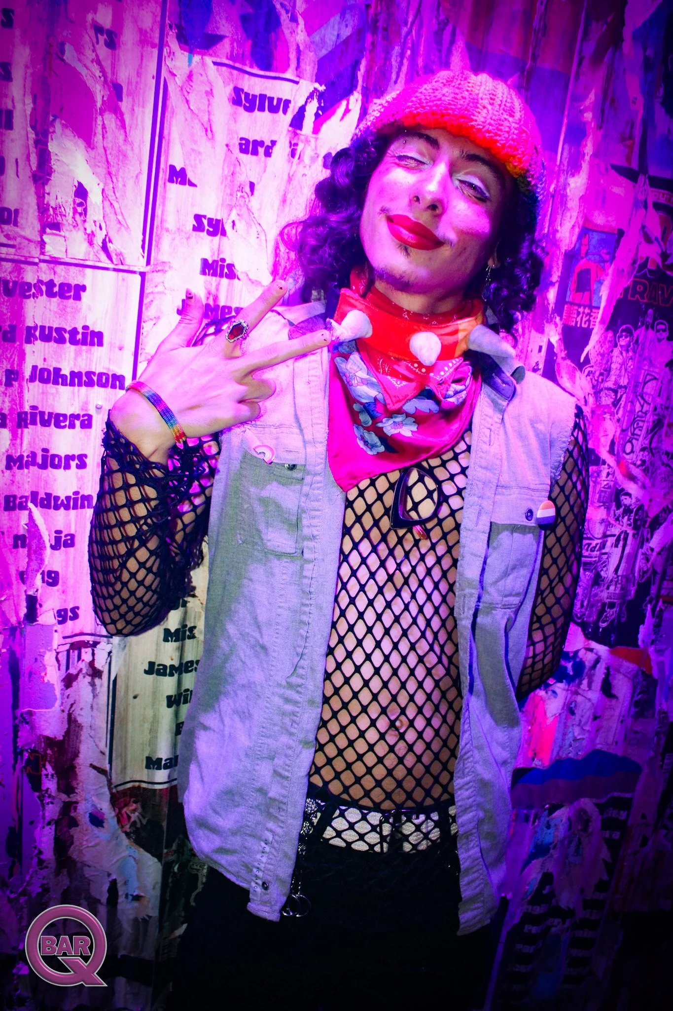 A person with curly hair, red lipstick, and wearing a red knit cap, fishnet top, and denim vest, making a peace sign in front of a torn paper wall with various posters and stickers, illuminated by pink and purple lighting.