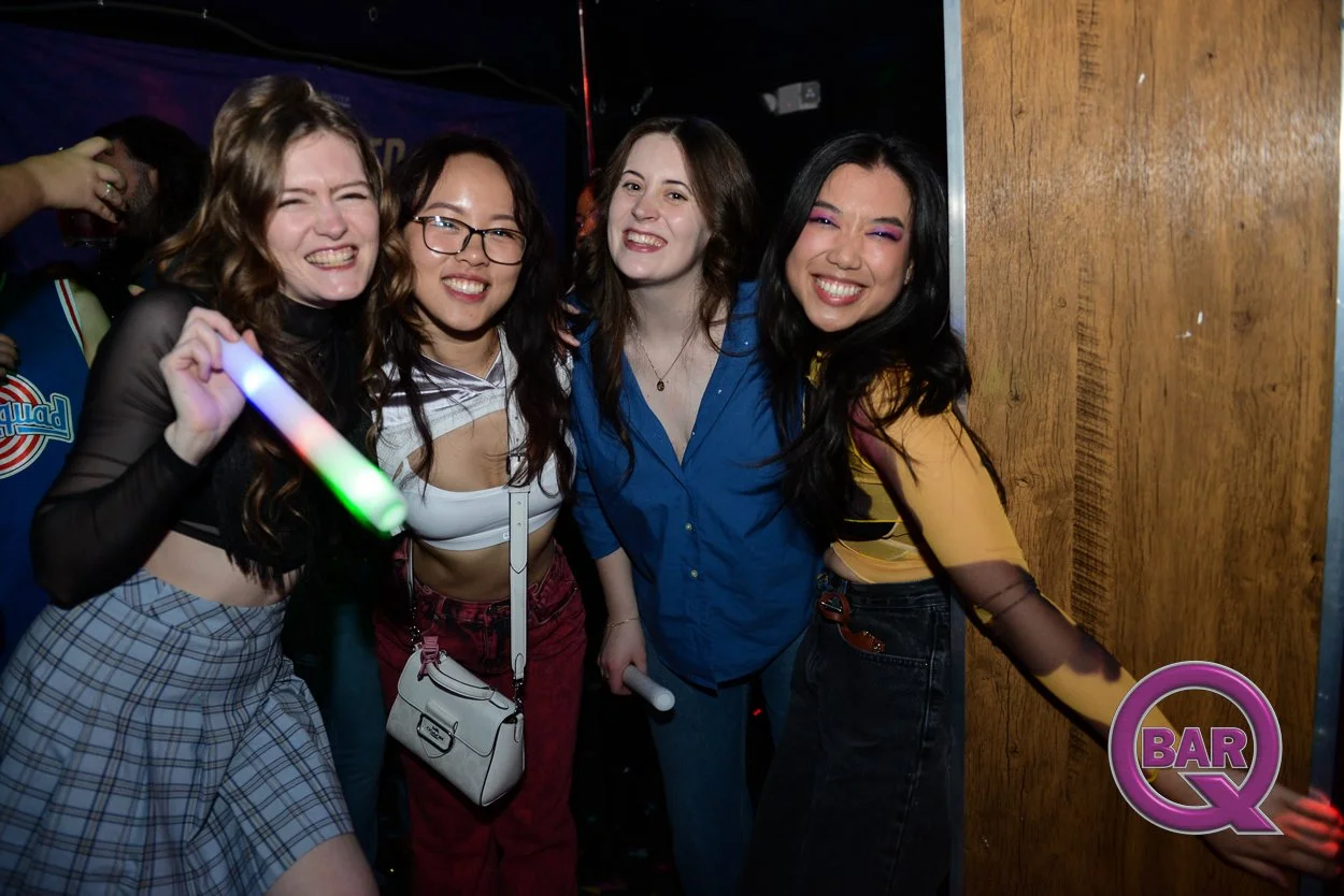 Four young women smiling and dancing at a bar or club with a wooden wall in the background. One woman is holding a glowing light stick, and they all appear to be happy and enjoying themselves.
