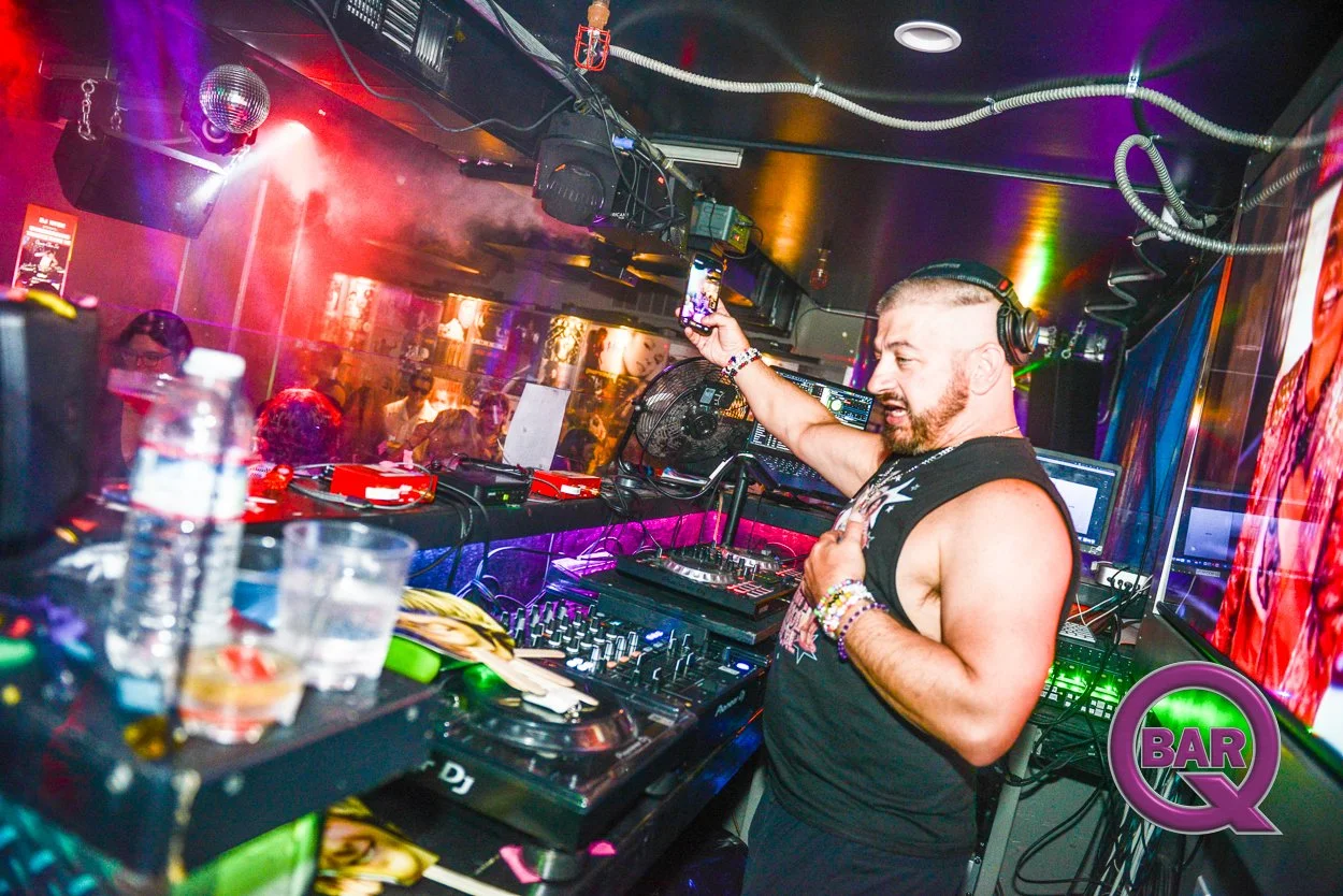 A DJ performing at a nightclub, wearing headphones and raising his phone in front of a crowd. Colorful lights and a large screen are visible in the background.