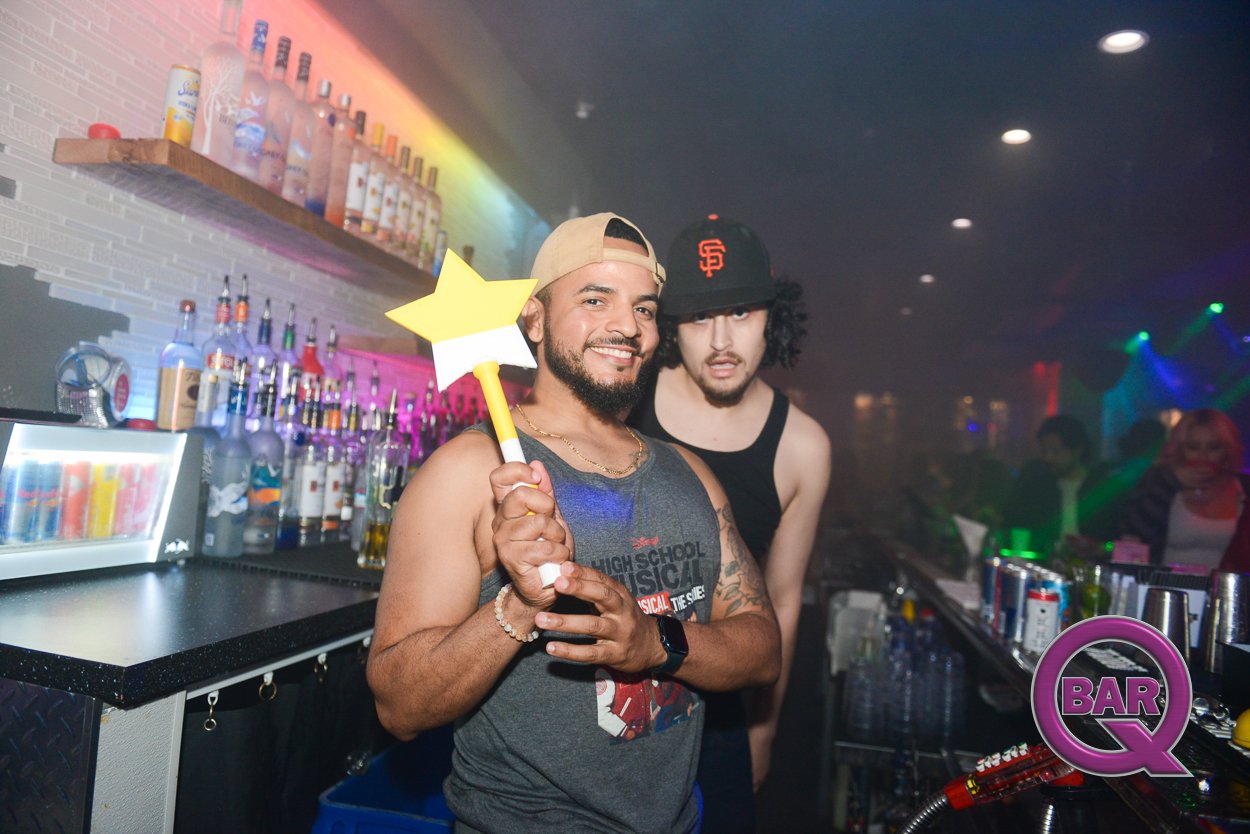 Two men behind a bar, one smiling and holding a star-shaped wand, the other standing next to him, both wearing tank tops and casual attire, with bottles of alcohol and bar supplies on the counter, and colorful lights in the background.