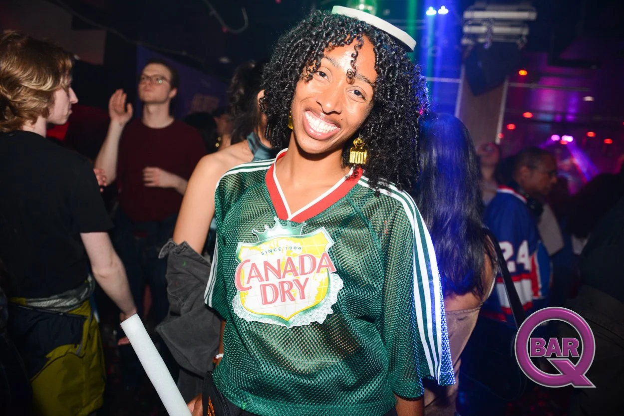 A woman with curly black hair and yellow earrings smiling at a party or nightclub, wearing a green Canada Dry jersey and a white headband, with colorful lighting and other people in the background.