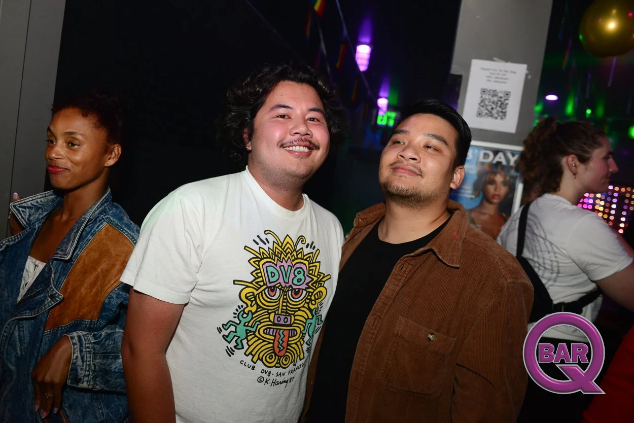 Two men smiling and posing for a photo in a bar or nightclub with other people, colorful lights, and a logo that says 'BAR Q' in the background.
