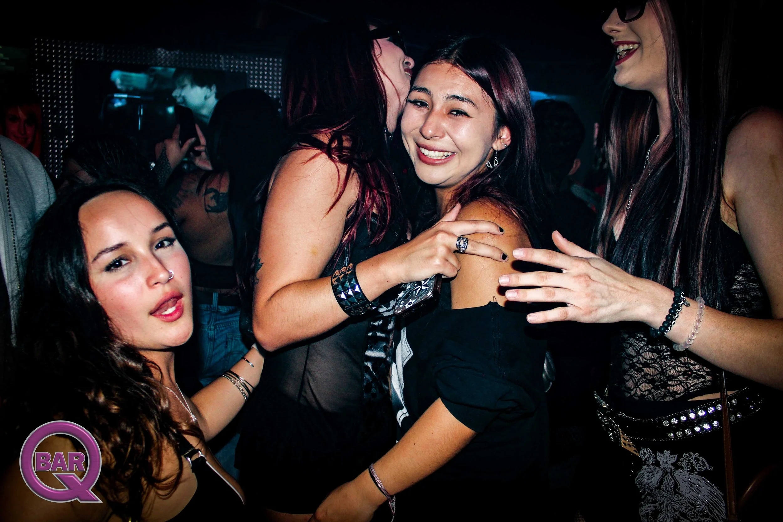 Group of young women dancing and having fun at a nightclub or bar, with one woman being kissed on the cheek by another woman.