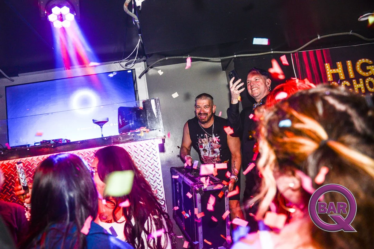 People dancing and celebrating at a DJ event with pink and purple confetti flying in the air. Two men are smiling at the front of the crowd, one holding up a phone. A DJ setup with a drink on the counter is visible in the background, with colorful li